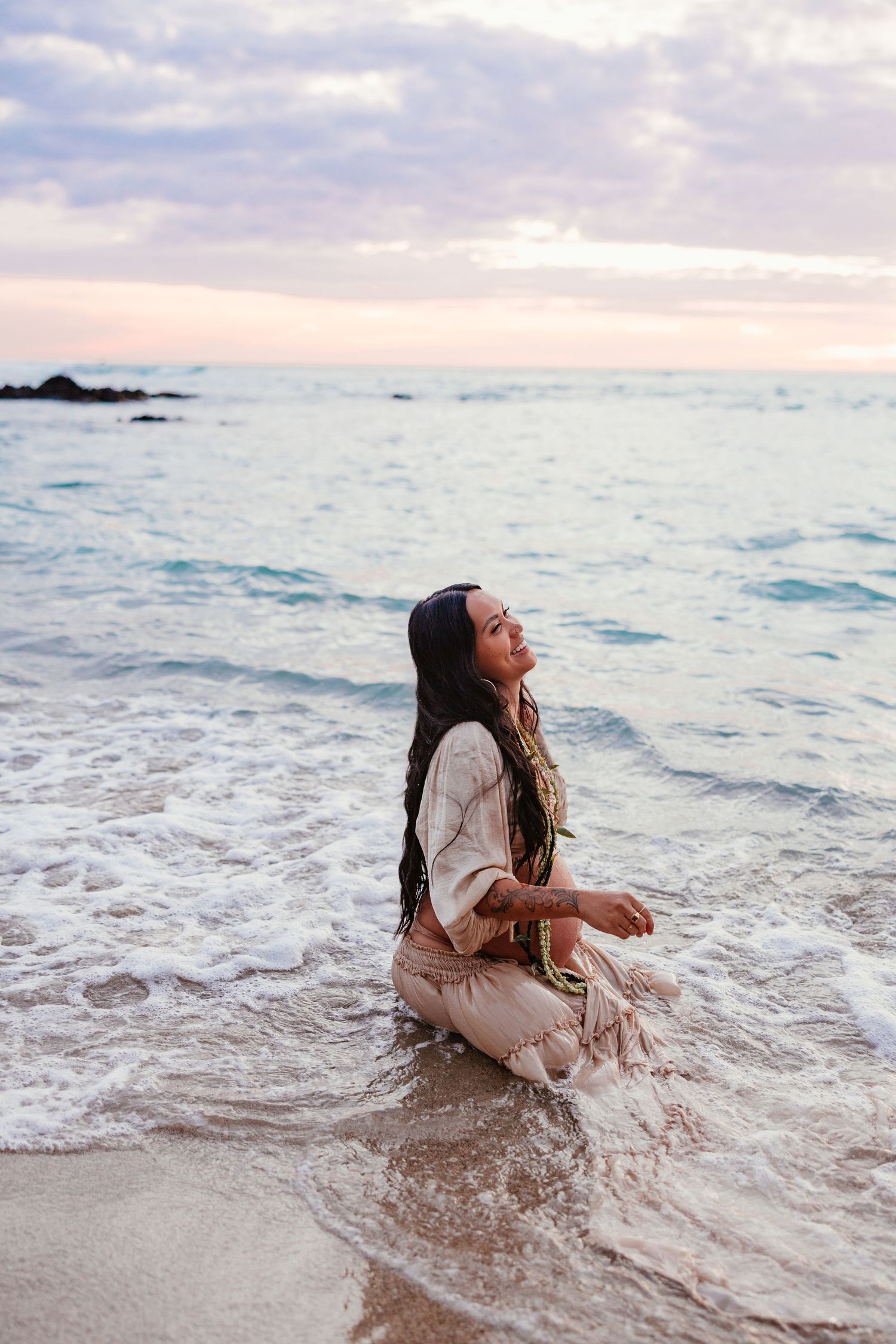 Woman in bohemian outfit sitting peacefully in ocean waves on a sandy beach at sunset.