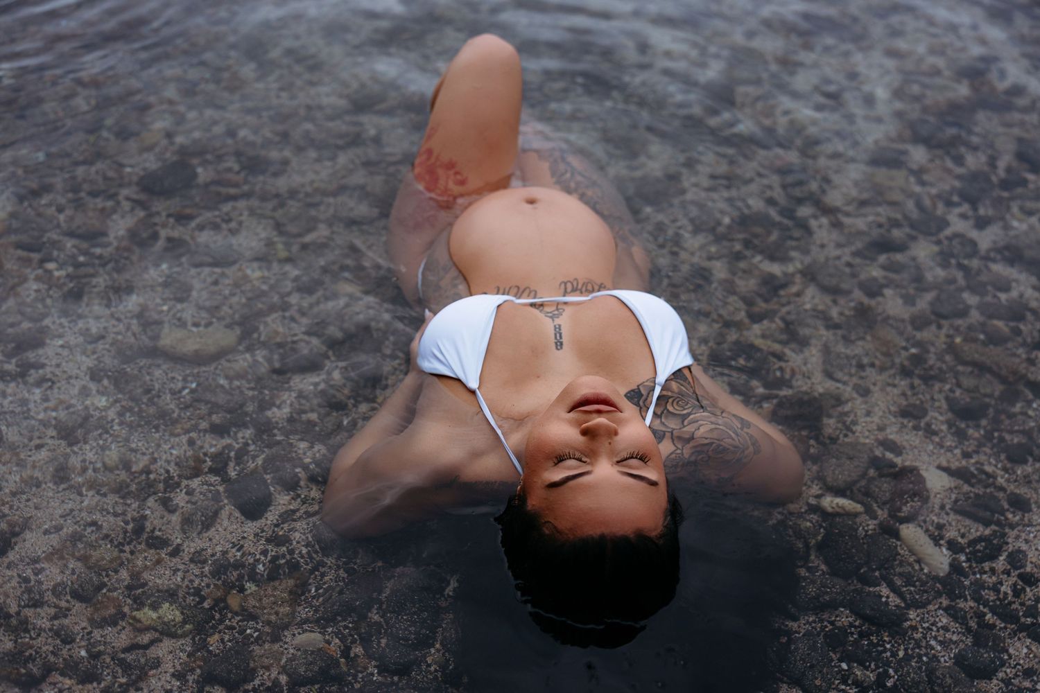 Woman in white bikini floating in dark water with eyes closed and peaceful expression.