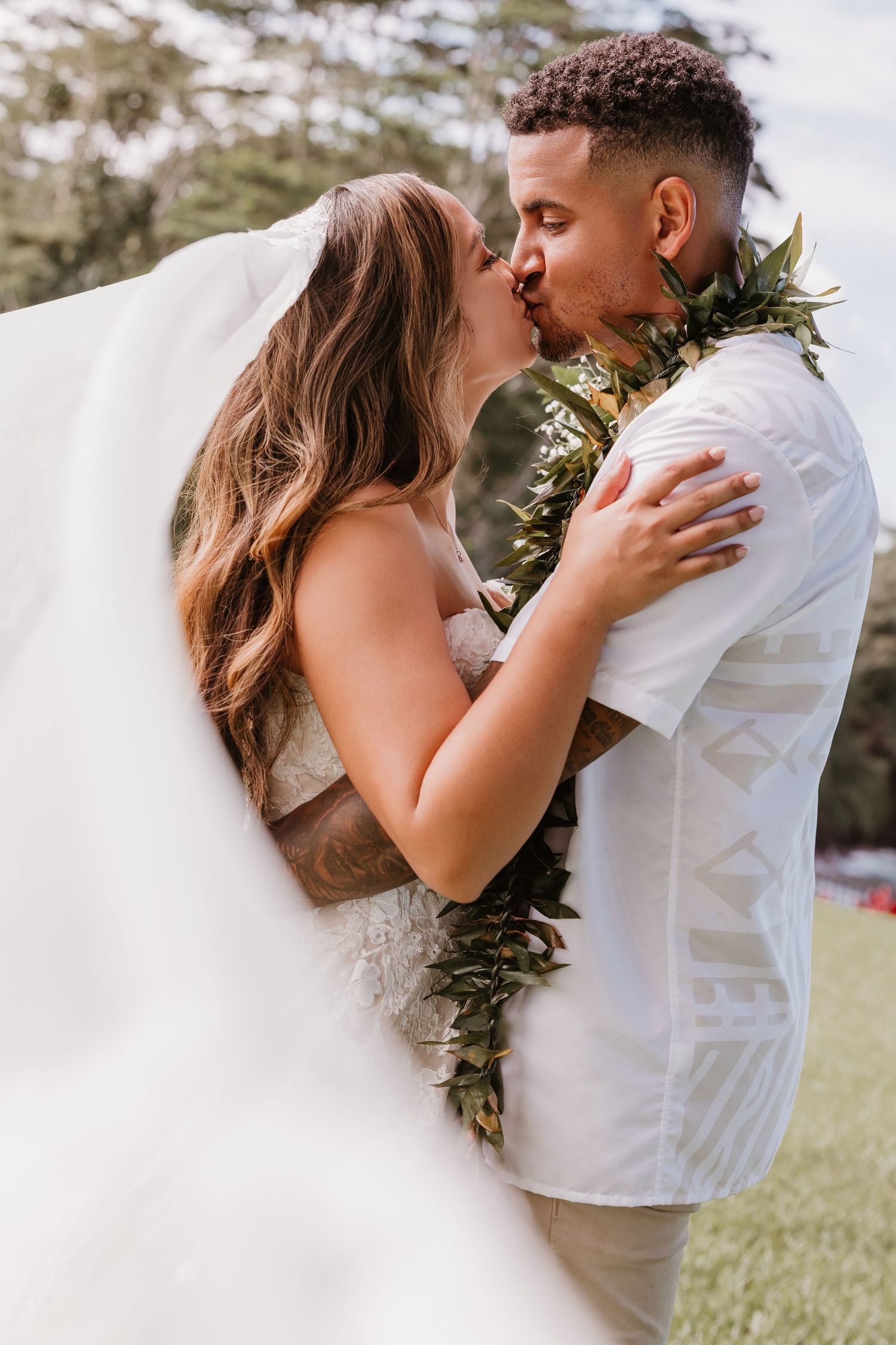 Bride and groom sharing a kiss outdoors, groom wearing a Hawaiian lei and white shirt at a tropical wedding.