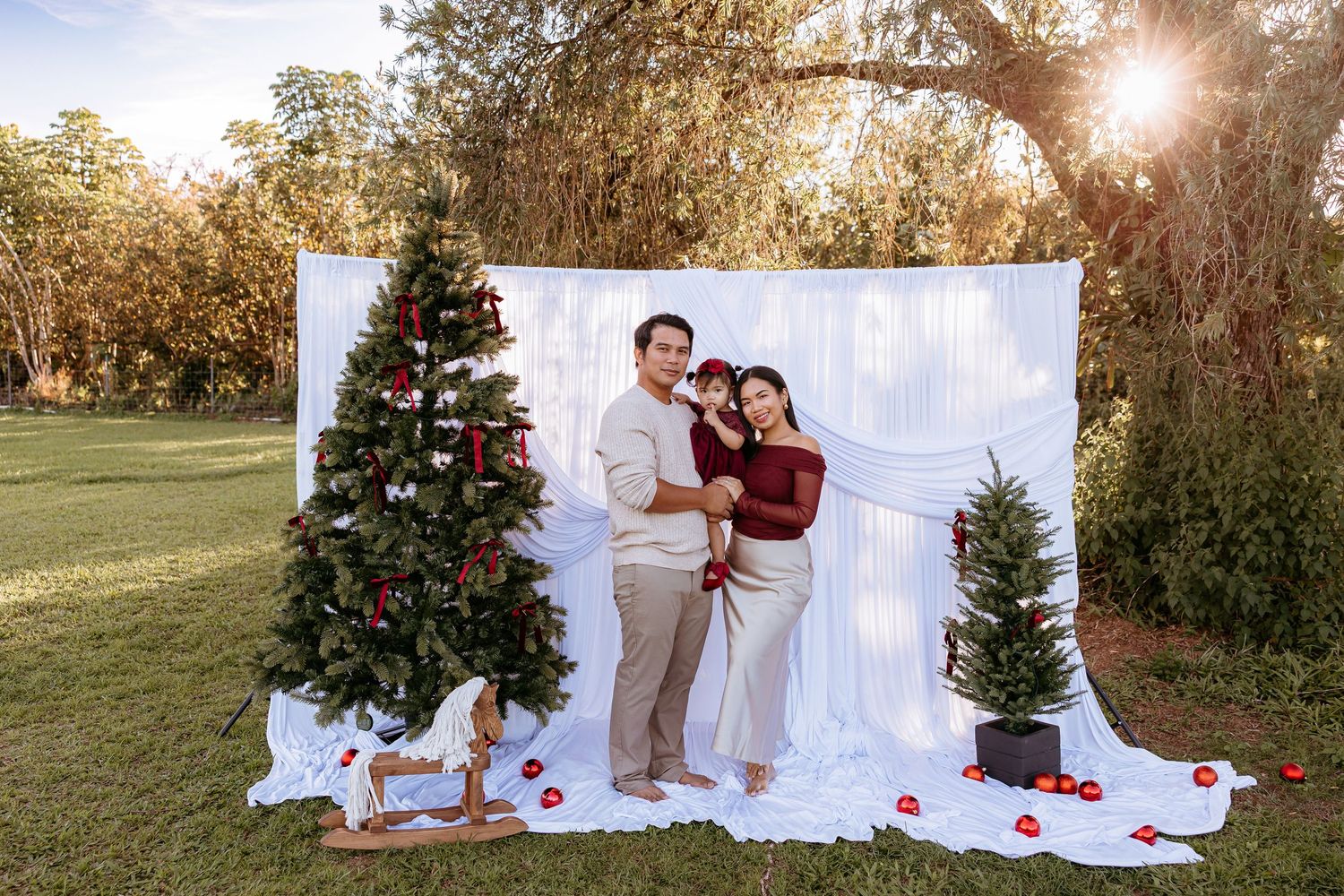 Holiday photo session outdoors with decorated Christmas trees and white backdrop against golden sunlight.