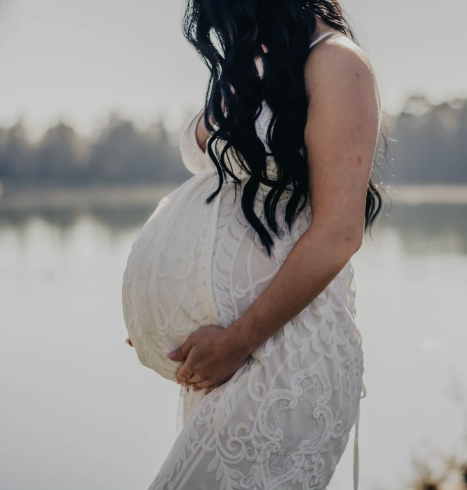 Silhouette of pregnant woman in lace dress standing by lake during maternity photoshoot.