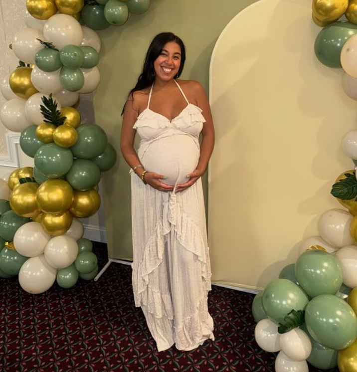 Expectant mother in white maxi dress poses at baby shower with mint green and gold balloon arch decorations.