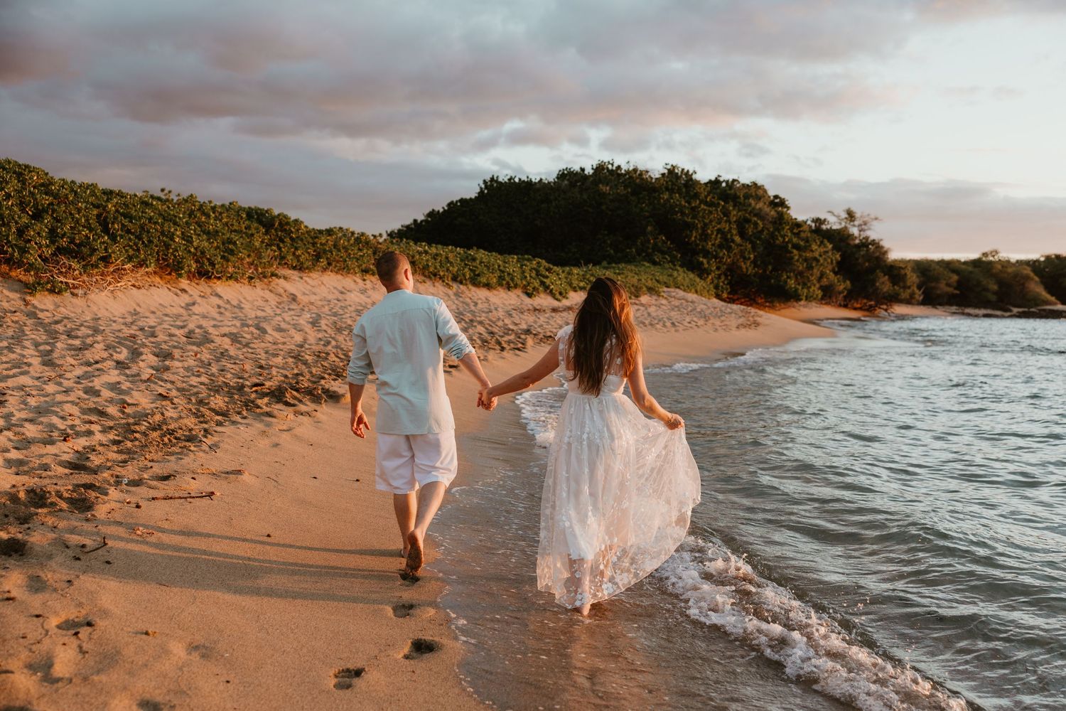 Couple walking hand in hand along a sandy beach at sunset.