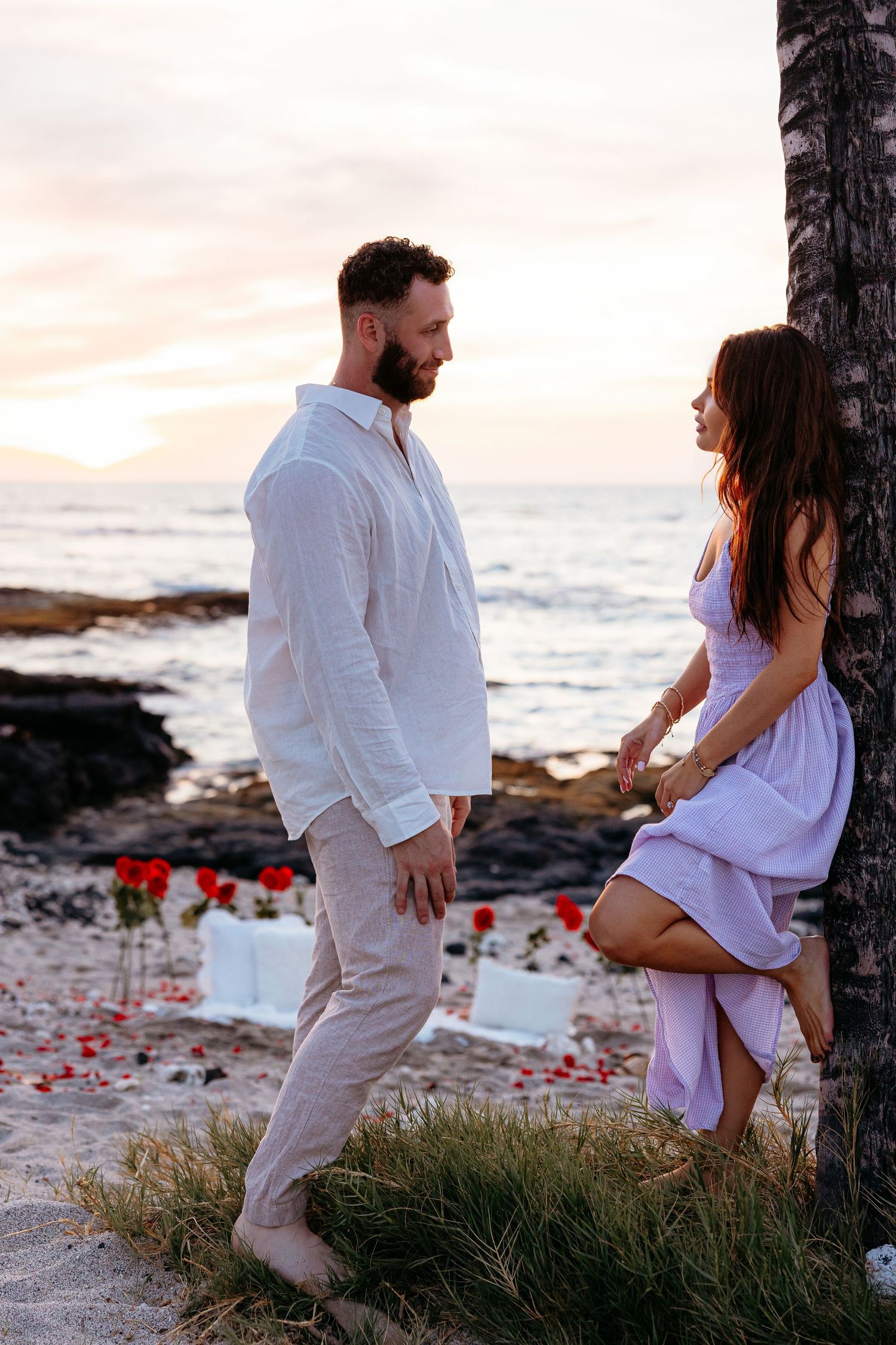 Couple holding hands on a rocky beach at sunset with red roses scattered on the ground behind them.