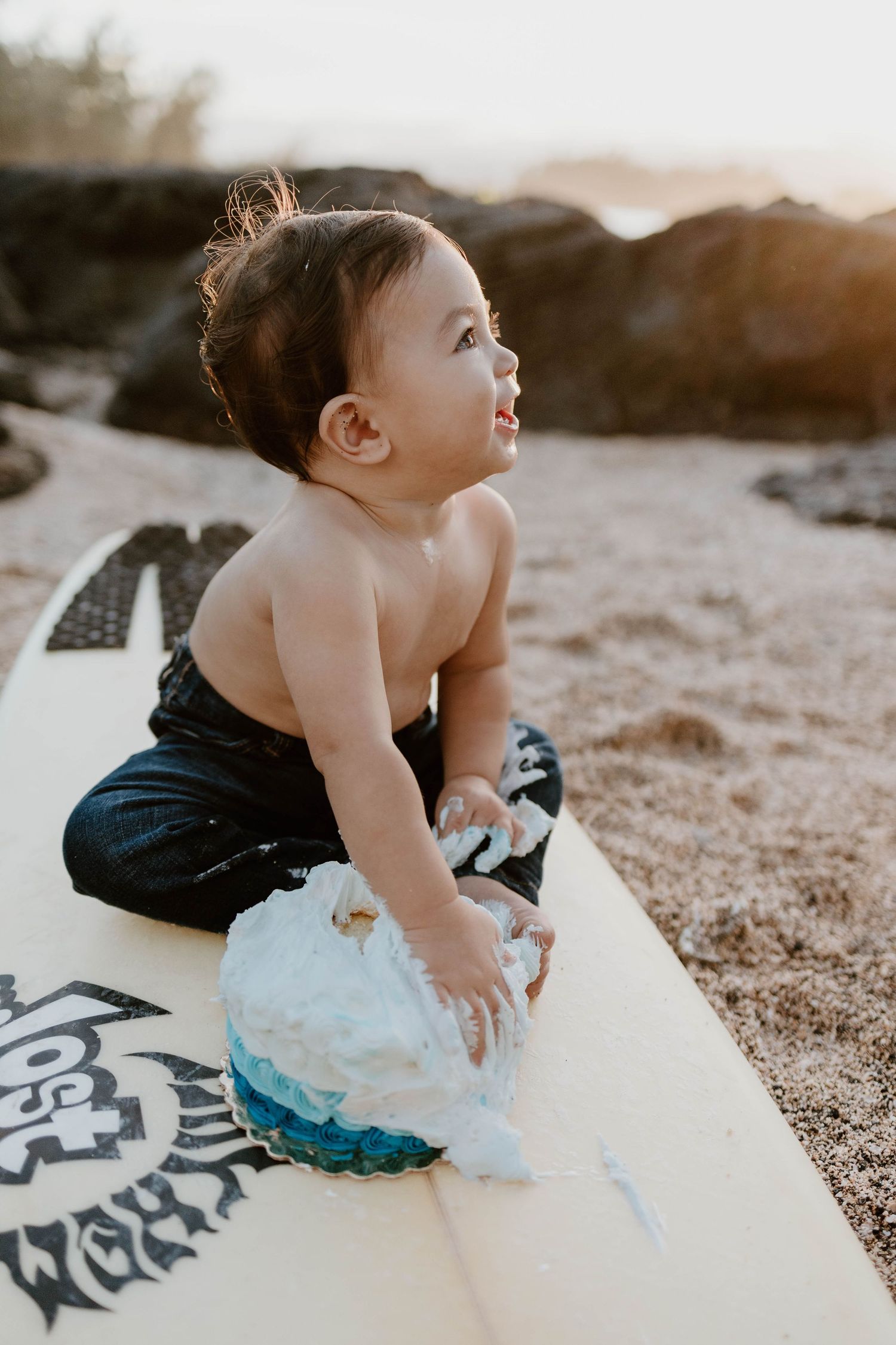 A baby sits on a surfboard at the beach during golden hour.