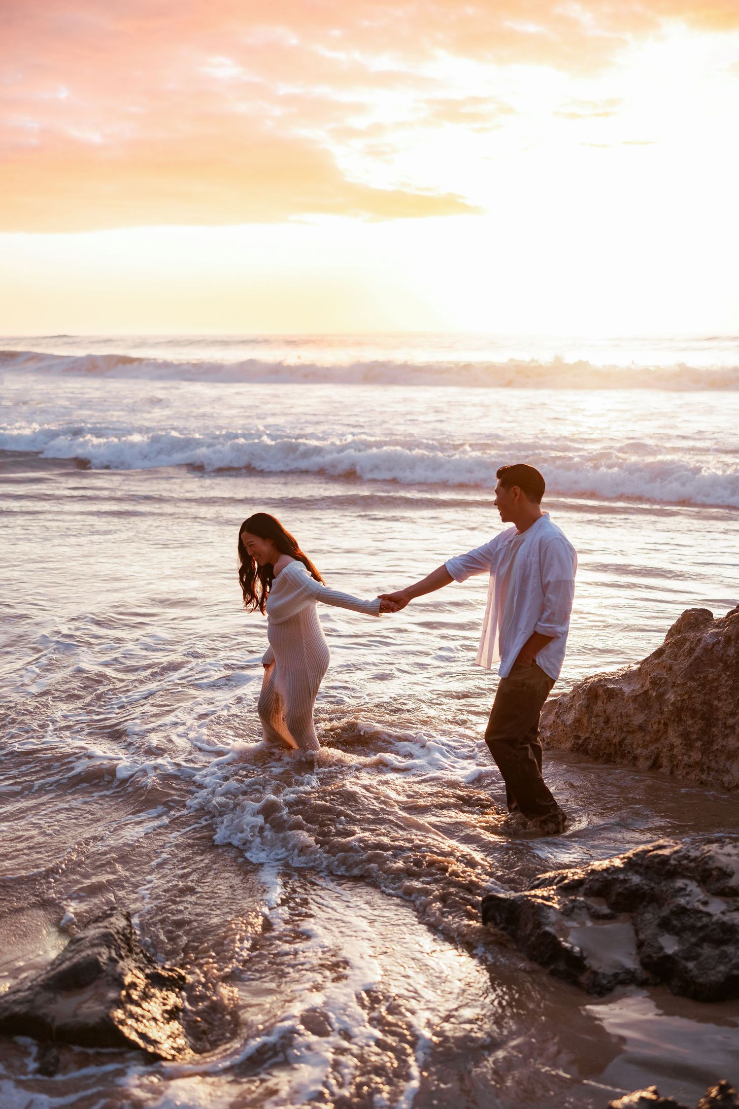 Pregnant couple holding hands walking in ocean waves at sunset on a rocky beach.
