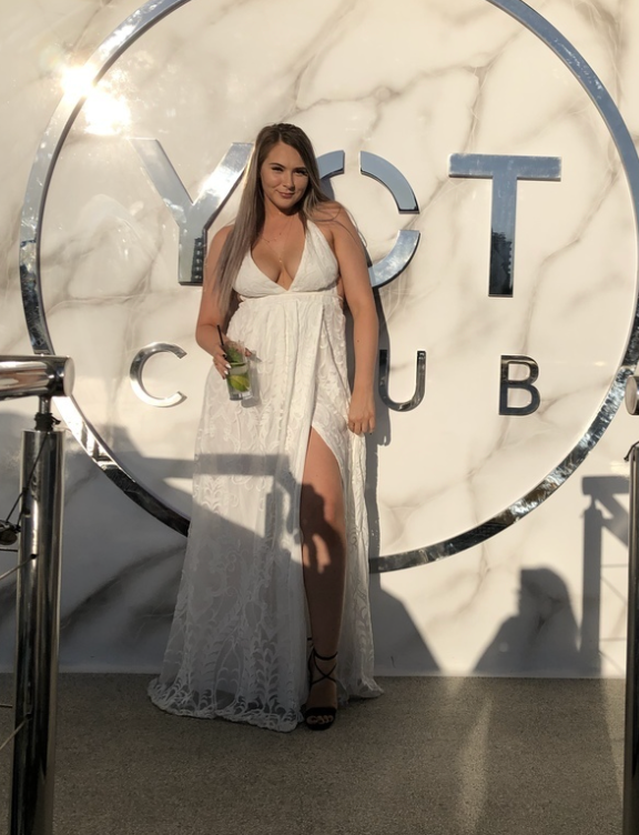 Woman in flowing white dress poses in front of illuminated YOT Club sign.