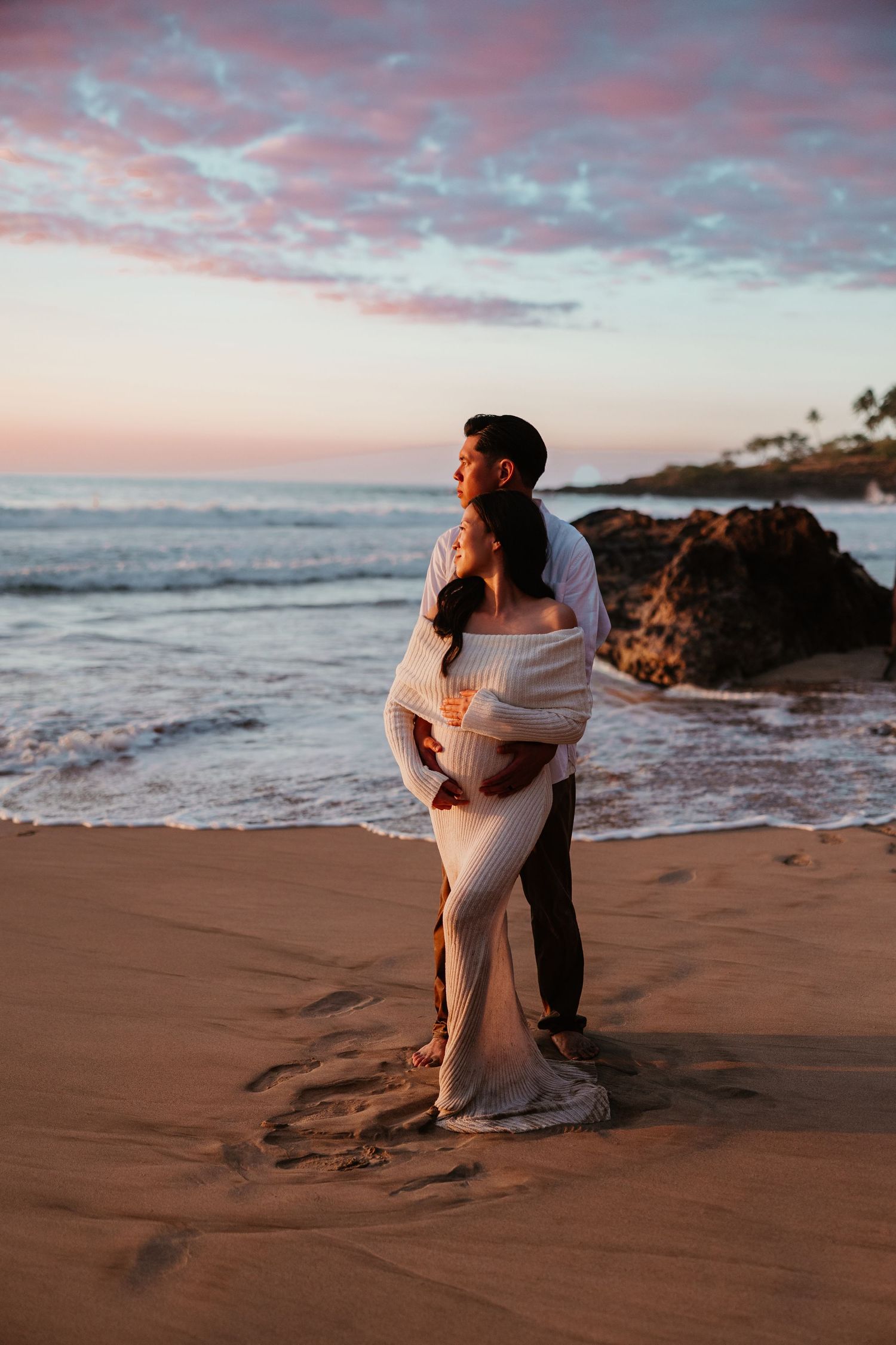 Couple embracing on a sandy beach at sunset with pink clouds and ocean waves in the background.