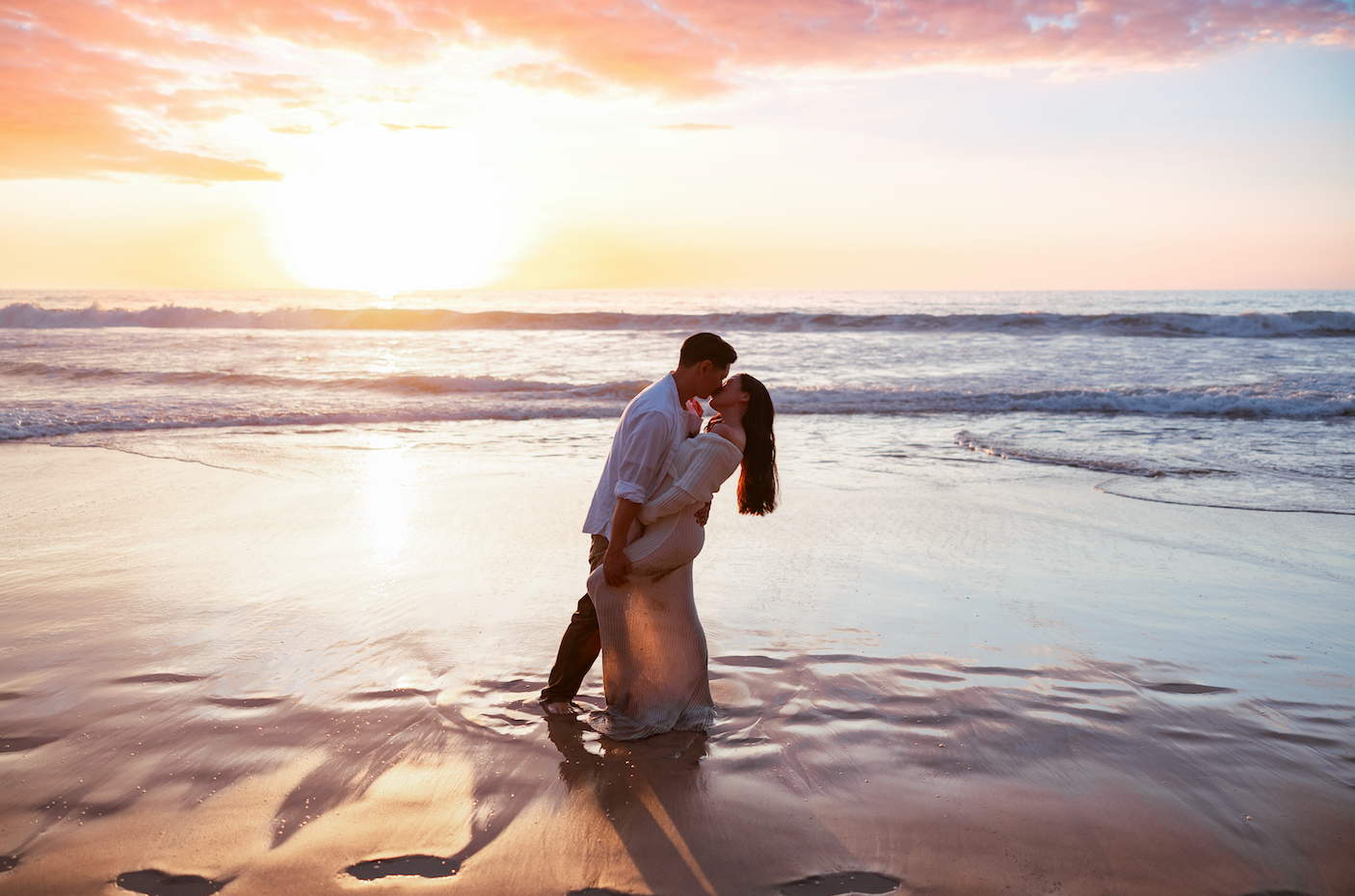 Couple sharing a romantic kiss on a sandy beach at sunset with golden waves and colorful sky in the background.