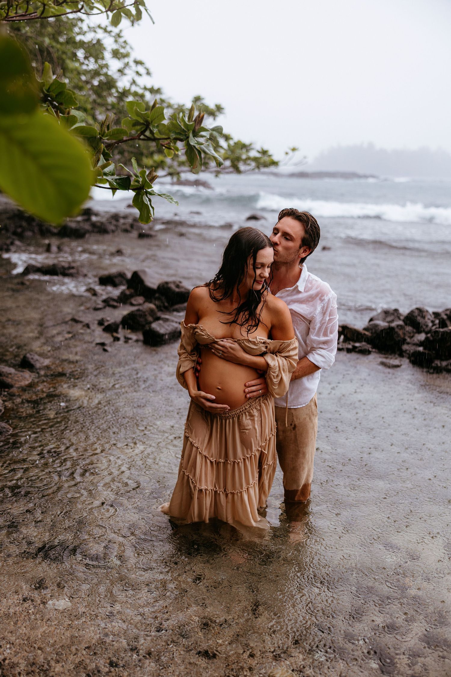 Couple sharing a tender maternity moment on a rocky Hawaiian beach, woman in flowing beige dress cradling baby bump.
