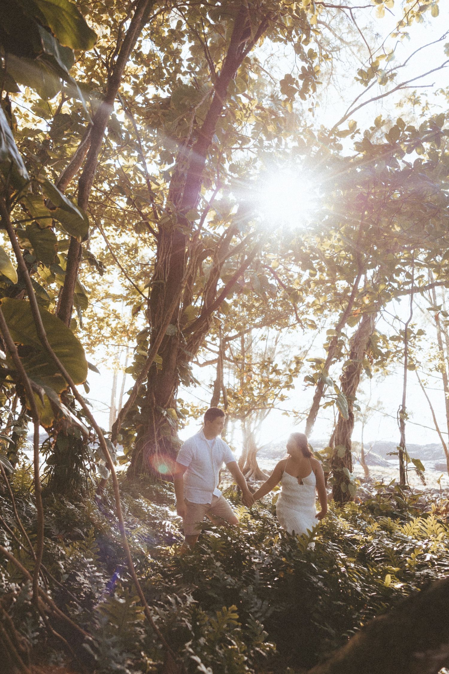 Couple in white attire holding hands while walking through sunlit tropical forest with golden lens flare.