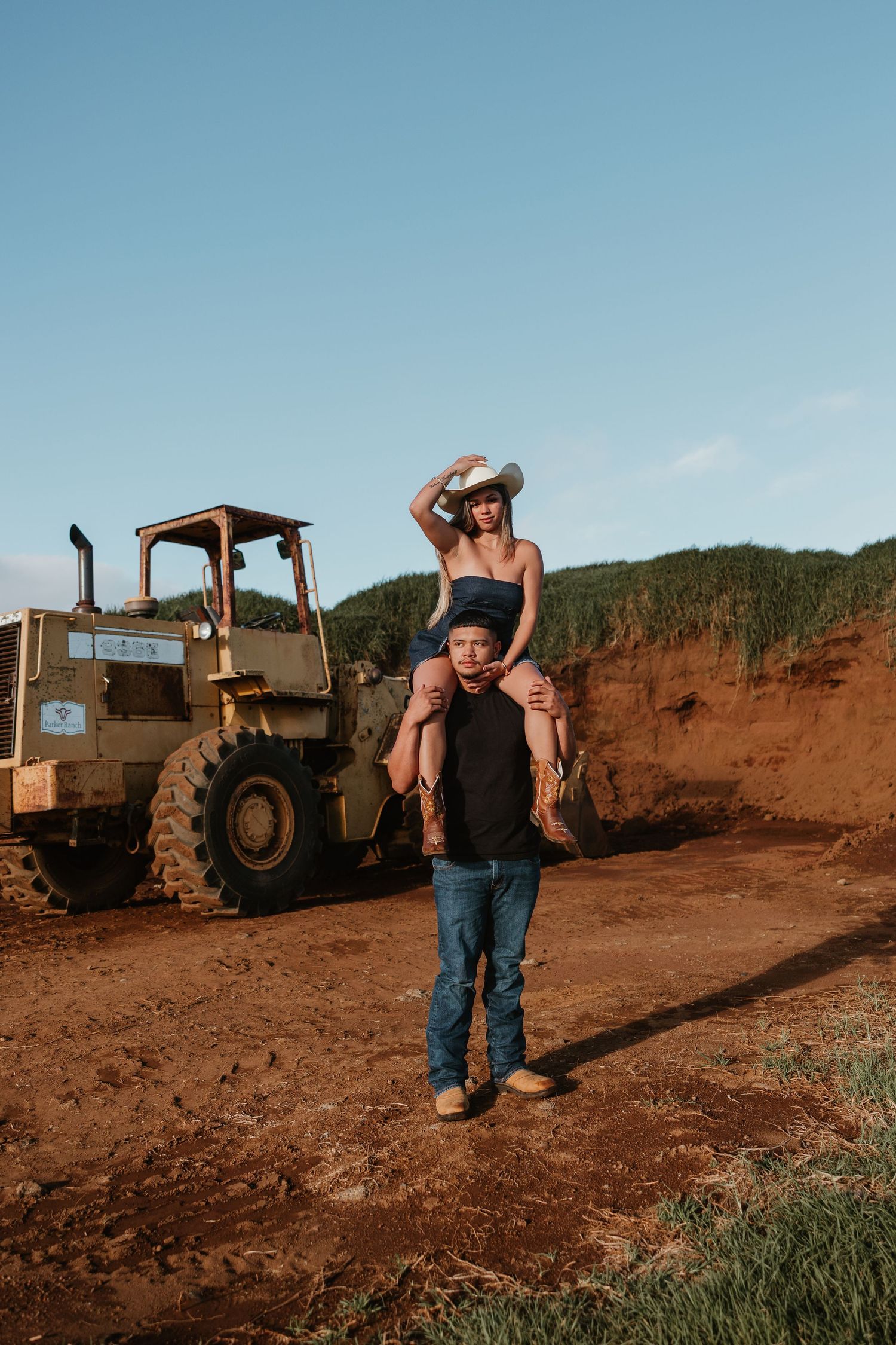 A woman in a cowboy hat sits on a man's shoulders near a vintage loader on a red dirt construction site.