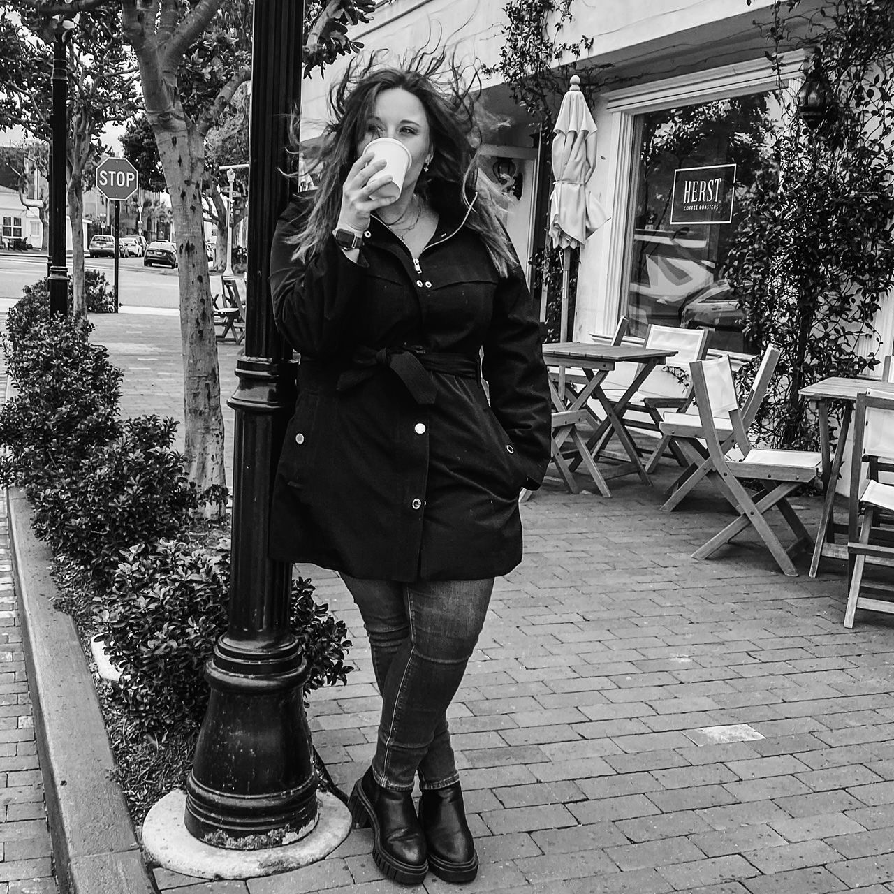 Black and white photo of woman drinking coffee on urban sidewalk outside cafe, wearing black coat.