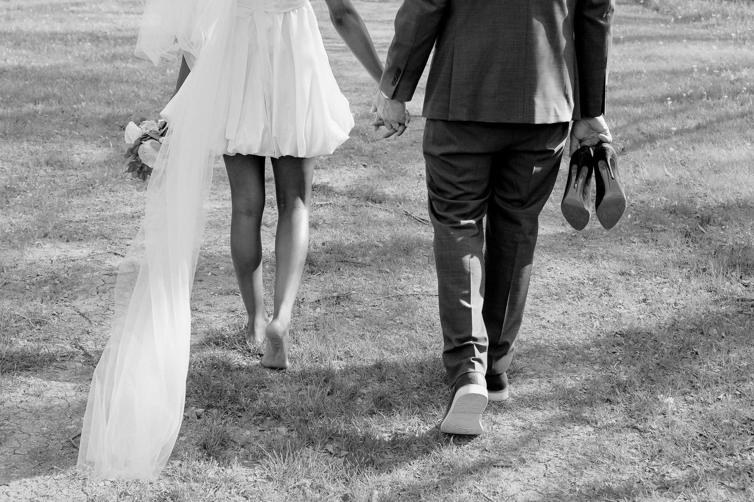 Black and white photo of bride and groom walking hand in hand across grass, groom carrying shoes.
