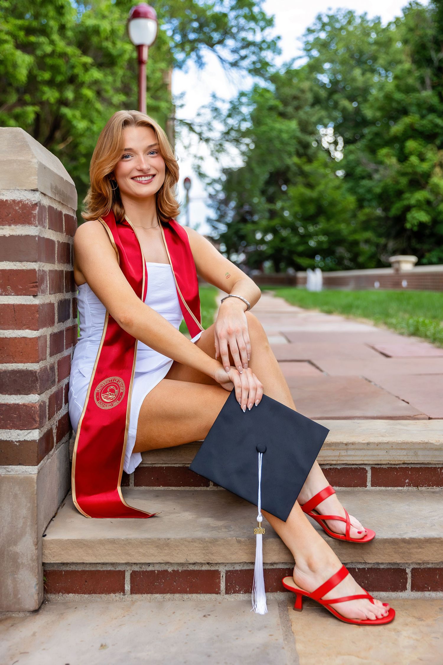 Smiling college graduate sits on brick steps wearing red sash and holding graduation cap.