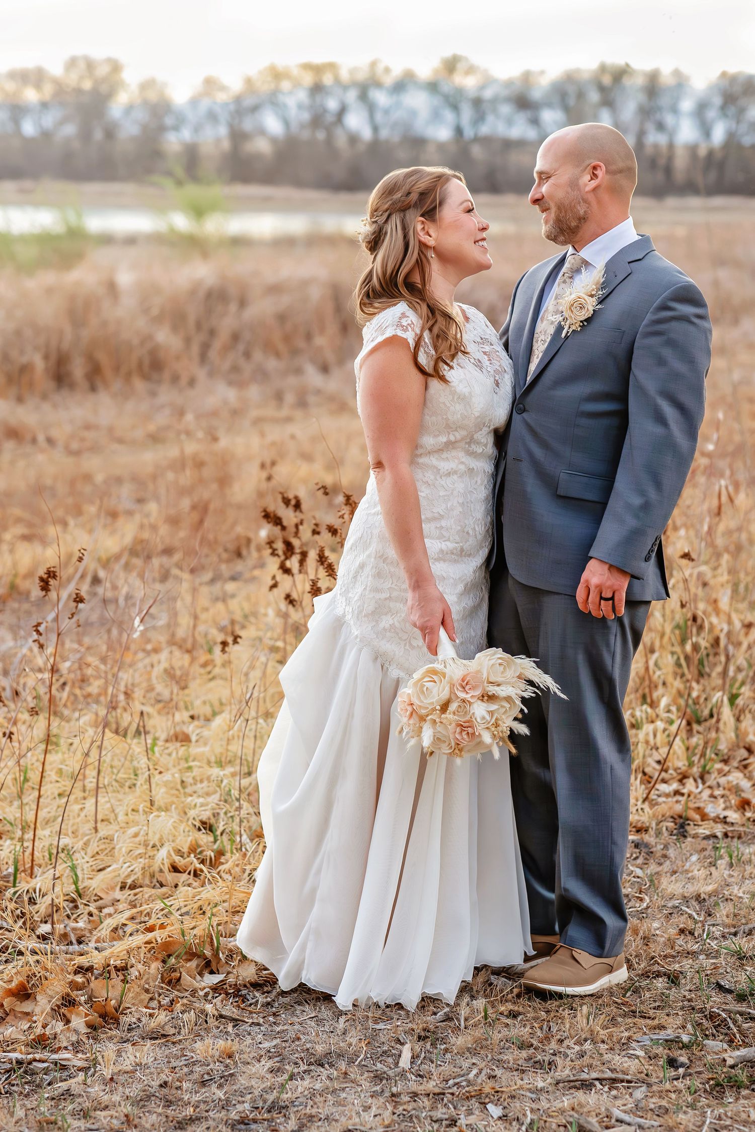 A happy bride and groom share a loving gaze in a dry autumn field with a lake backdrop.