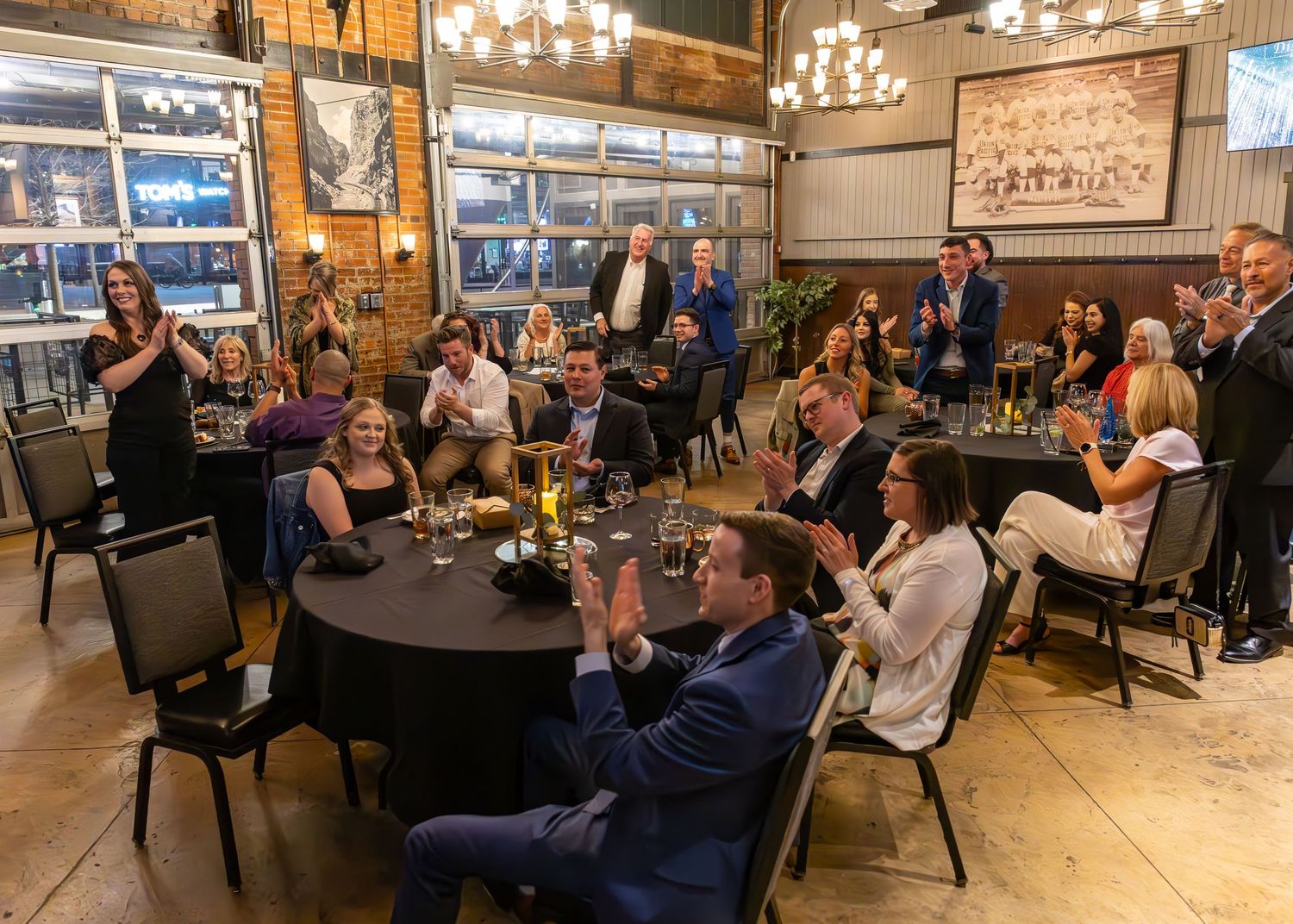 Guests applaud and celebrate at an elegant indoor dinner event with chandeliers and round tables.