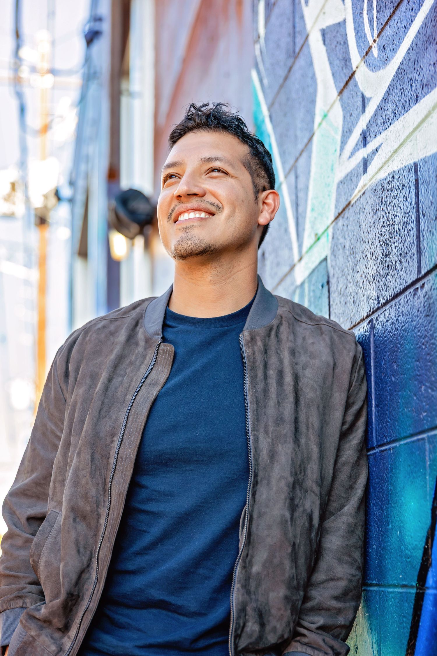 A smiling man in a brown bomber jacket leans against a colorful graffiti wall outdoors.