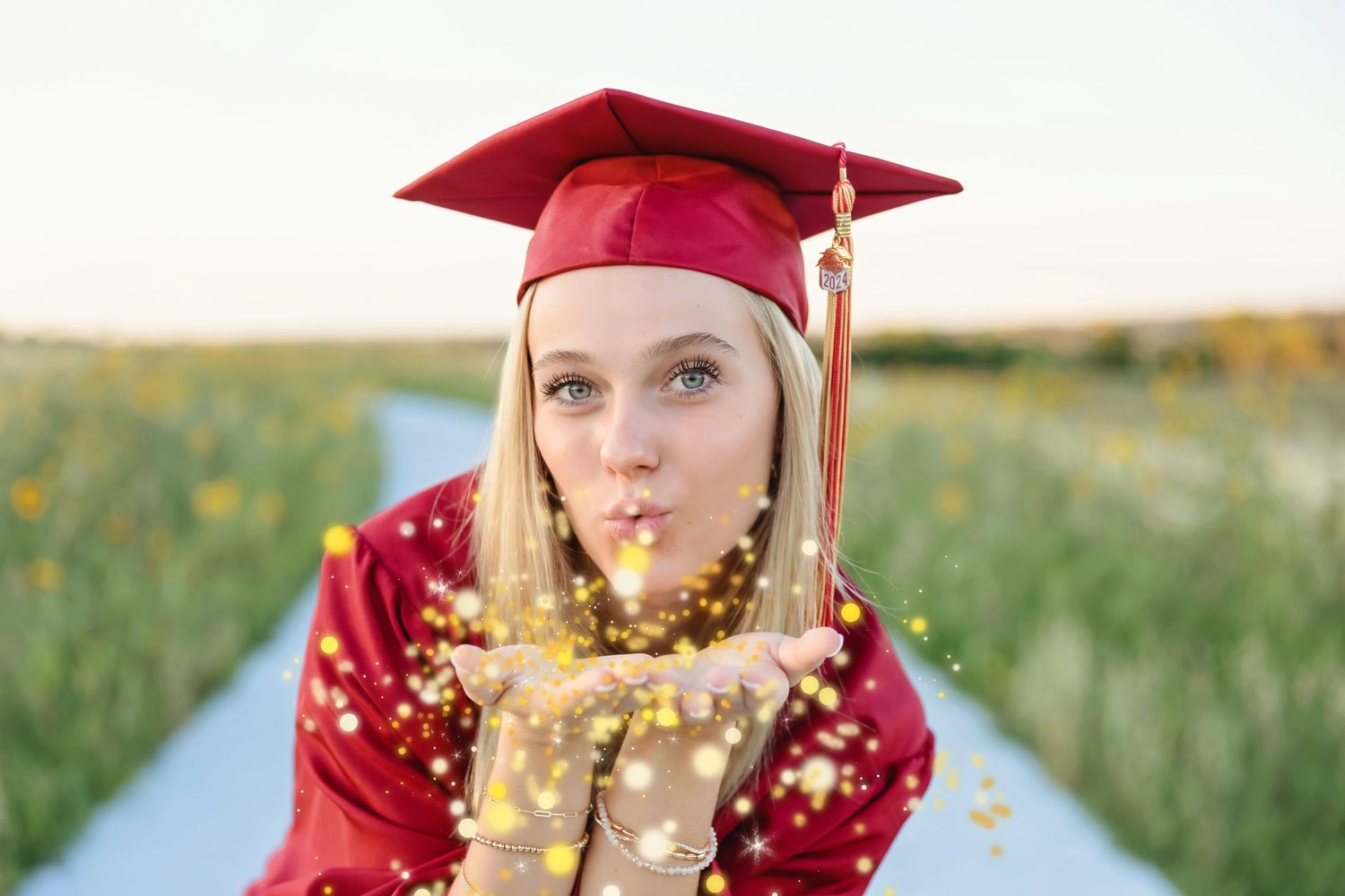 Blonde graduate in red cap and gown blowing gold glitter confetti in a sunny field.