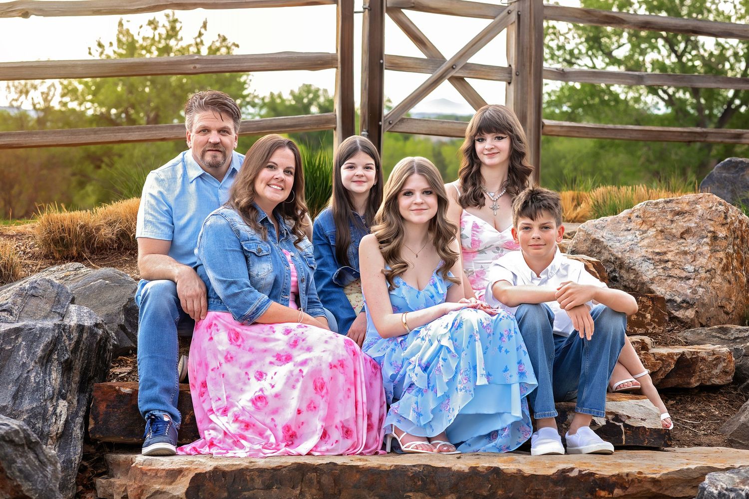 A smiling family of six poses on stone steps outdoors near a wooden gate at golden hour.