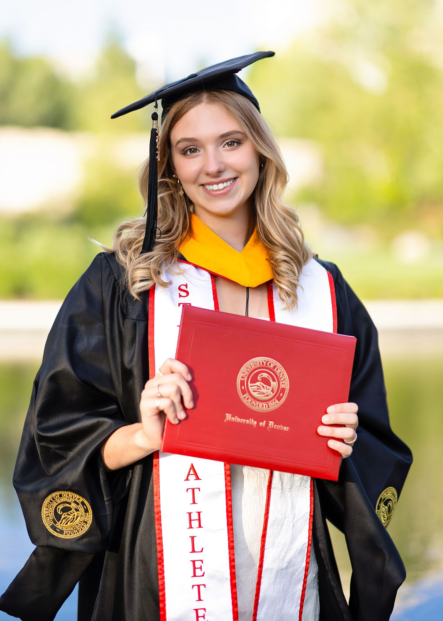 Smiling graduate in cap and gown holds University of Denver diploma outdoors near pond.