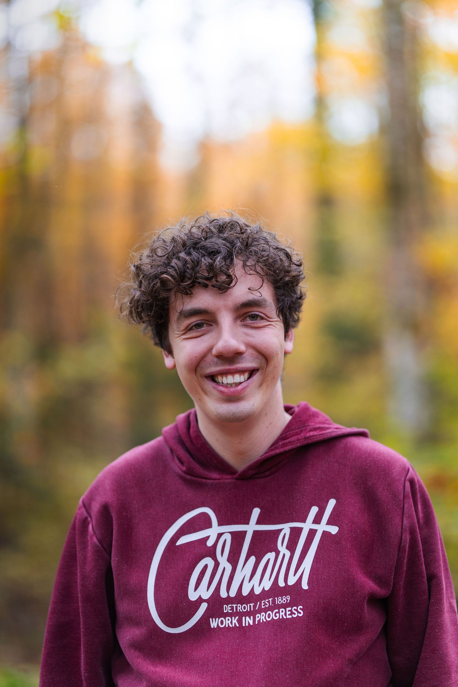 Young person in burgundy Carhartt hoodie smiling in autumn forest.
