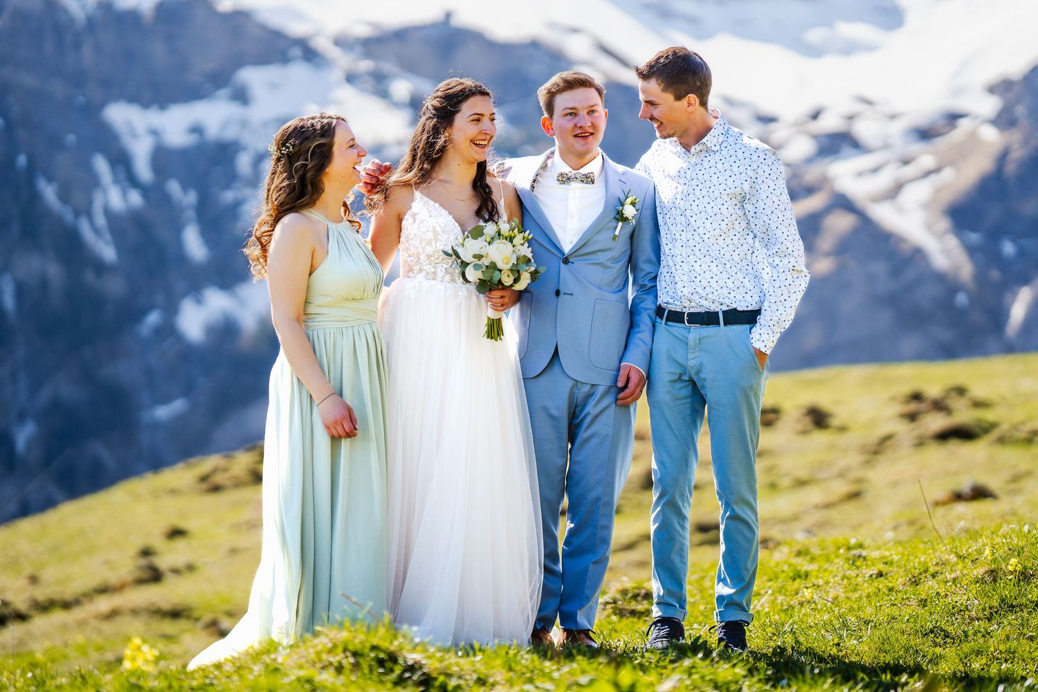 A wedding party poses together on a mountain meadow with snow-capped peaks in the background.