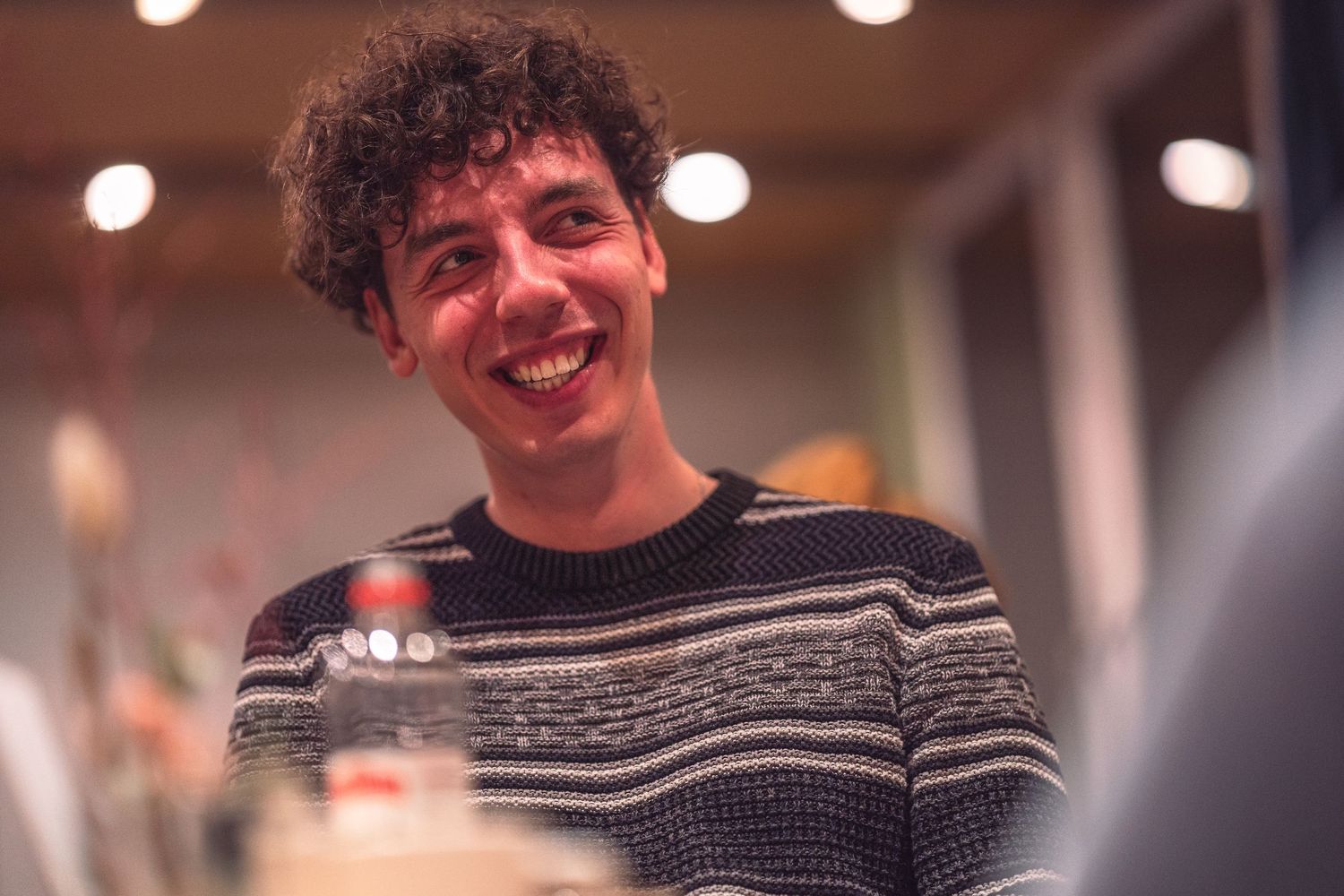 A candid shot of someone smiling while wearing a striped sweater under warm indoor lighting.