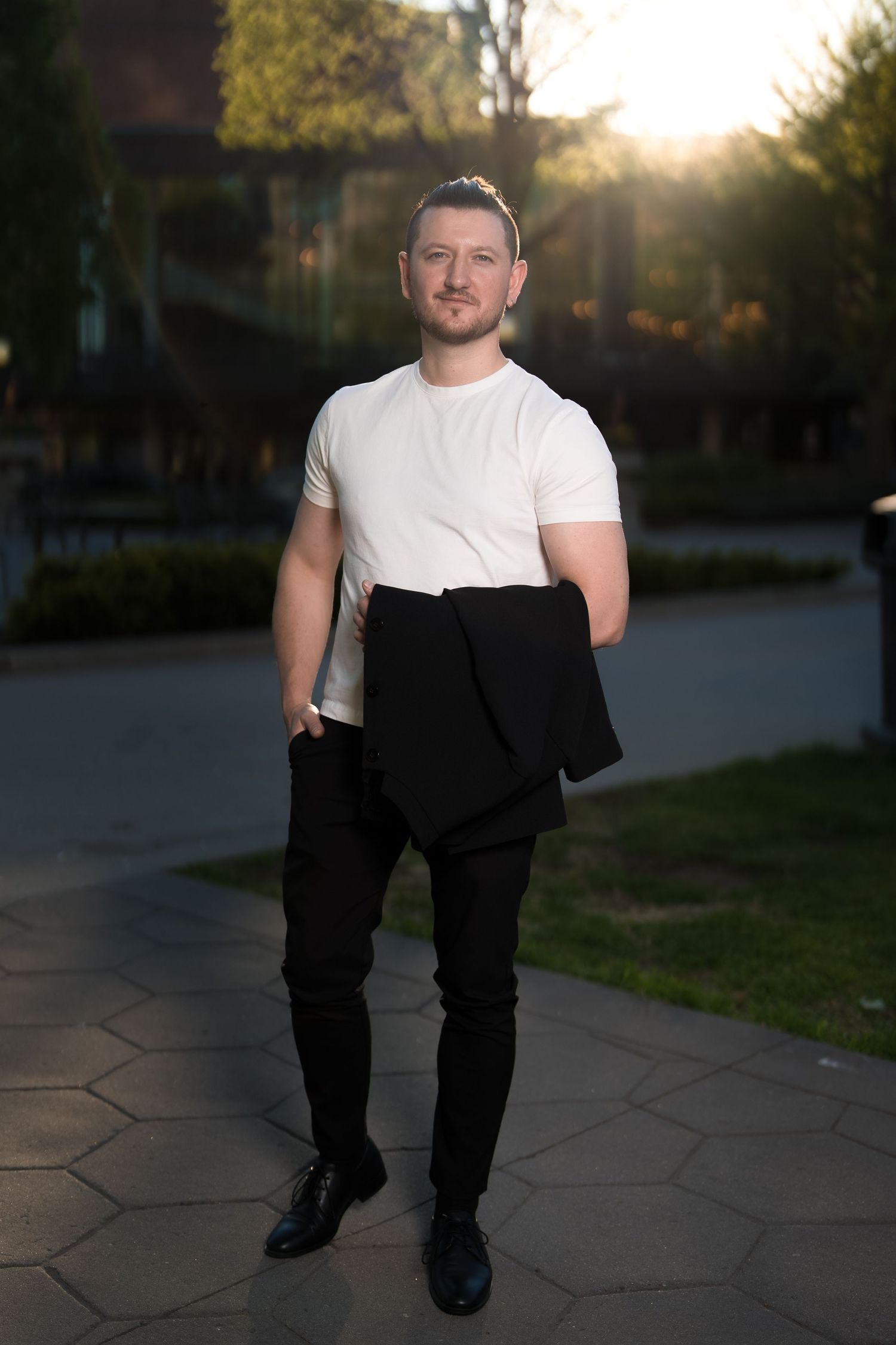 Man in white t-shirt and black pants holding jacket outdoors at sunset on a paved path.