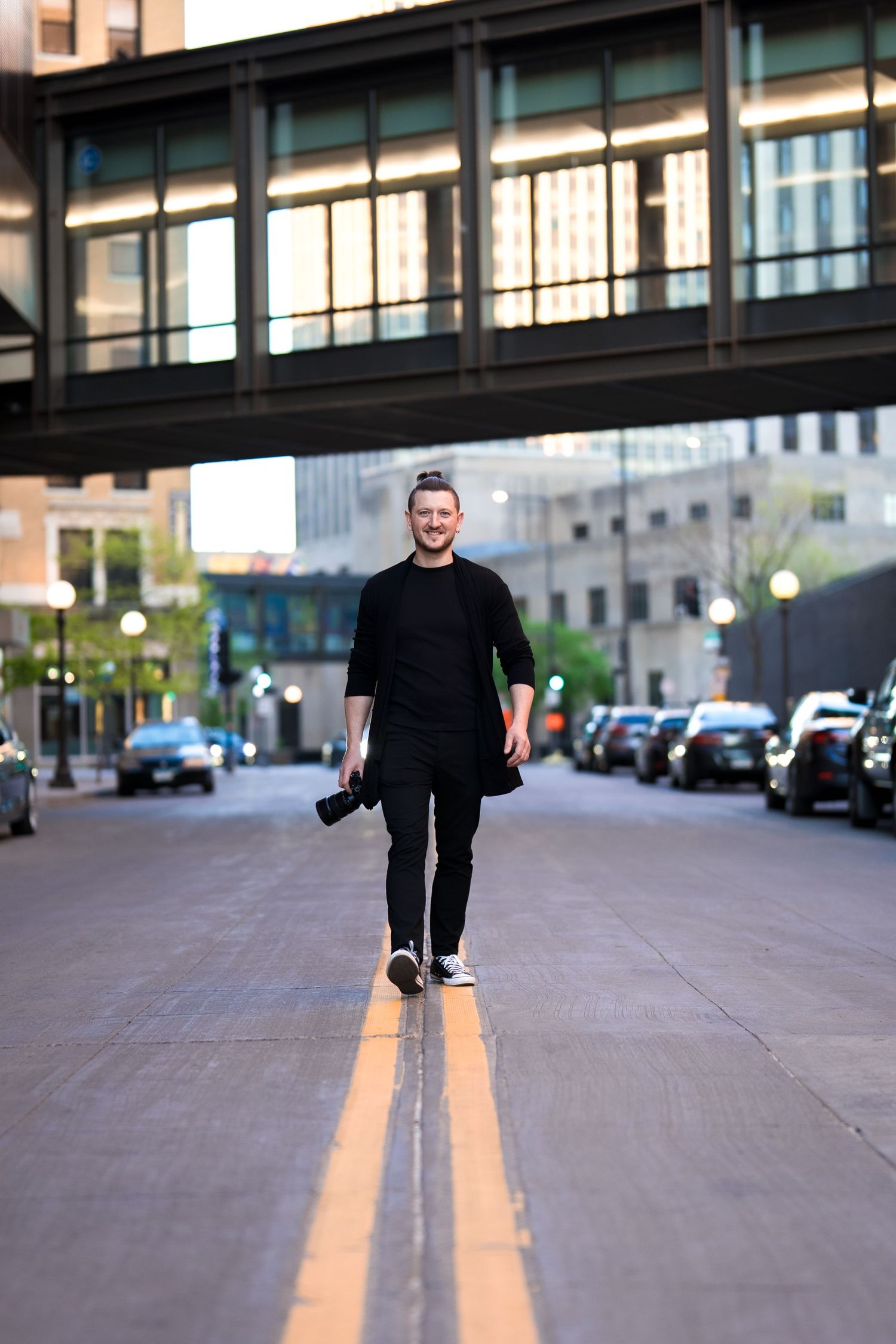A smiling man in all-black outfit walks down an urban street holding a camera, with city buildings and an elevated bridge behind him.