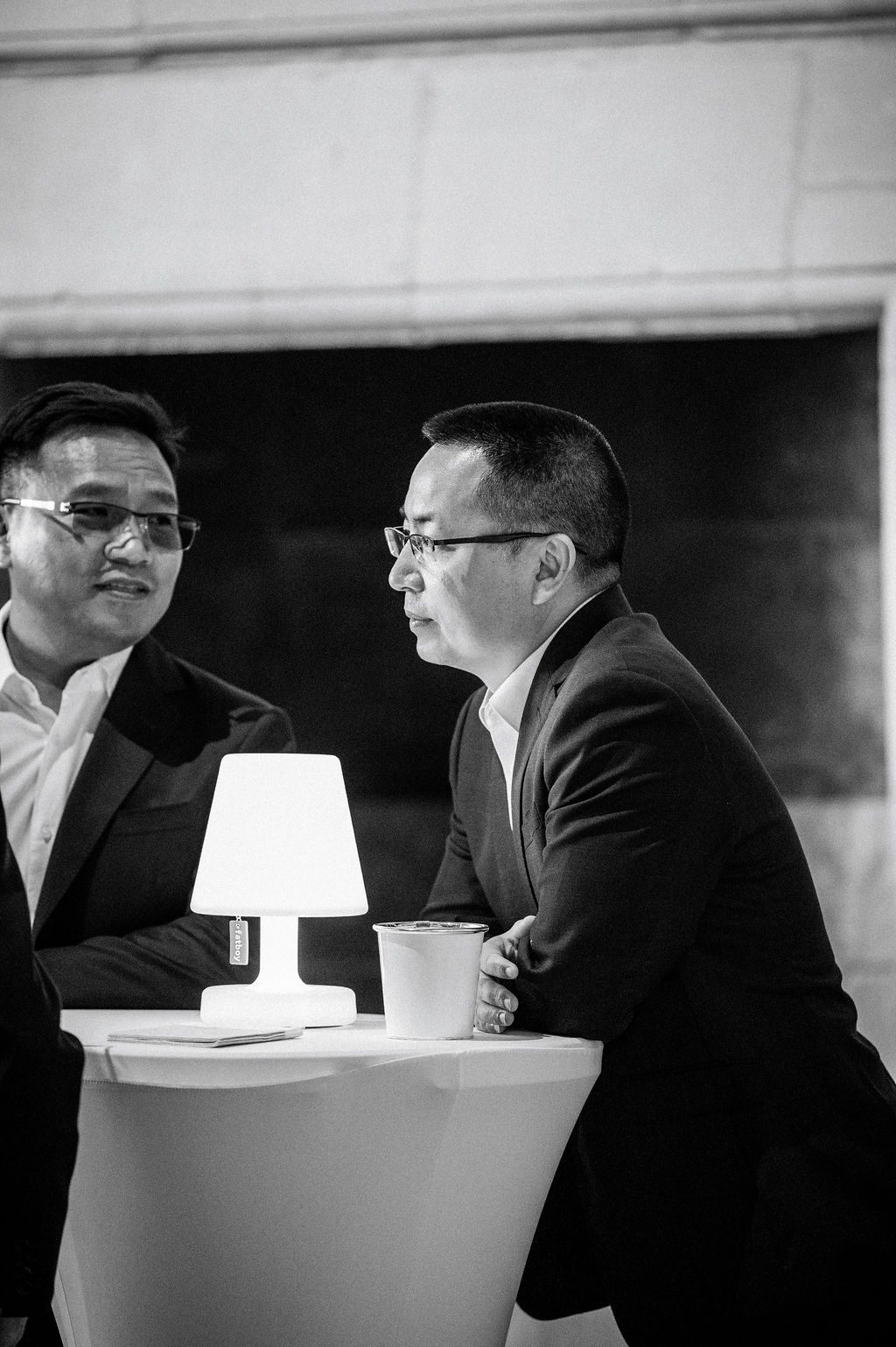 Deux dirigeants en costume discutent autour d’une lampe design sur table haute en intérieur.