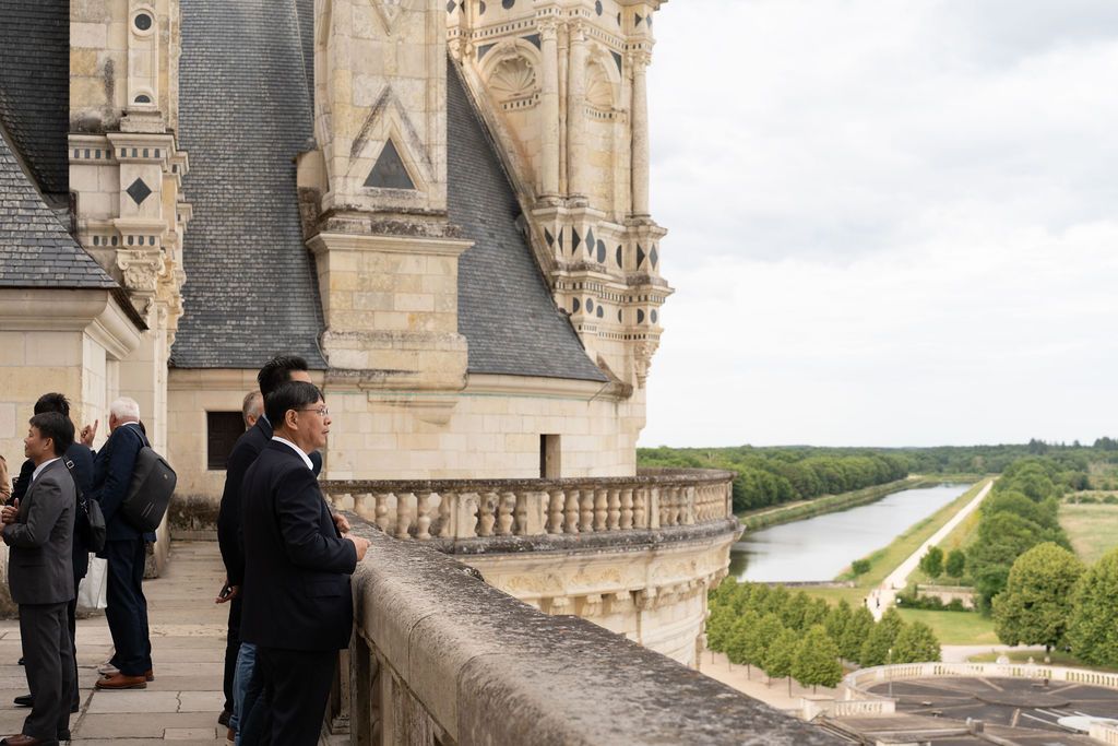 Groupe de décideurs échangeant lors d’un cocktail, vue partielle du château en arrière.