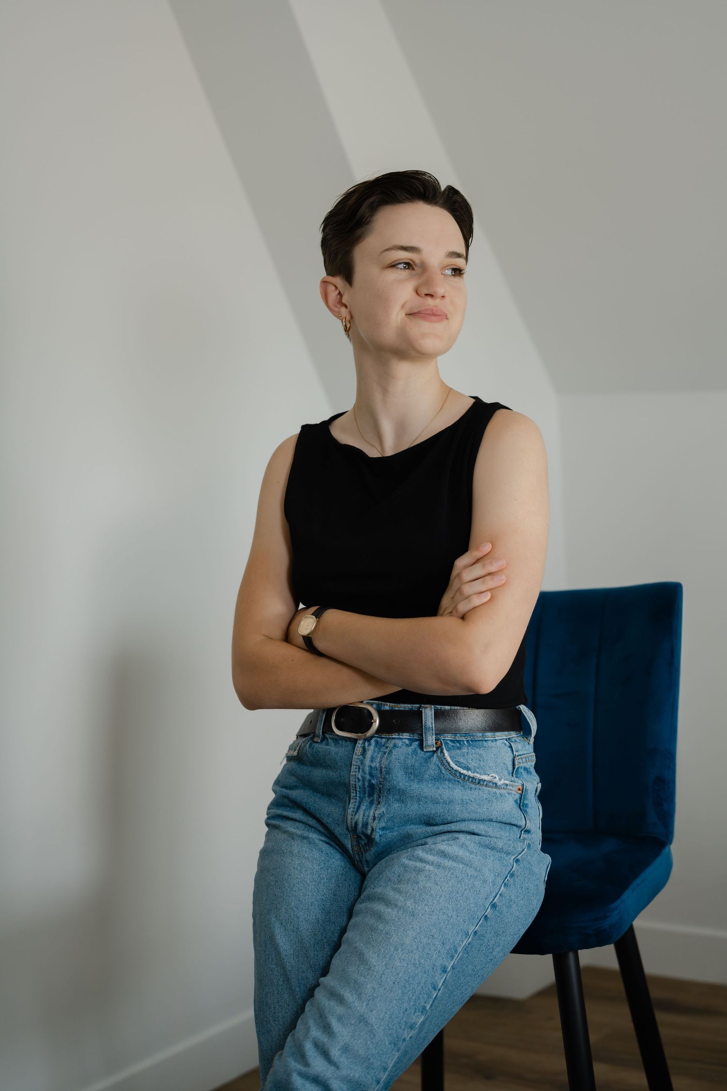 Person with short hair in black sleeveless top and jeans, arms crossed, leaning against blue chair in bright room.