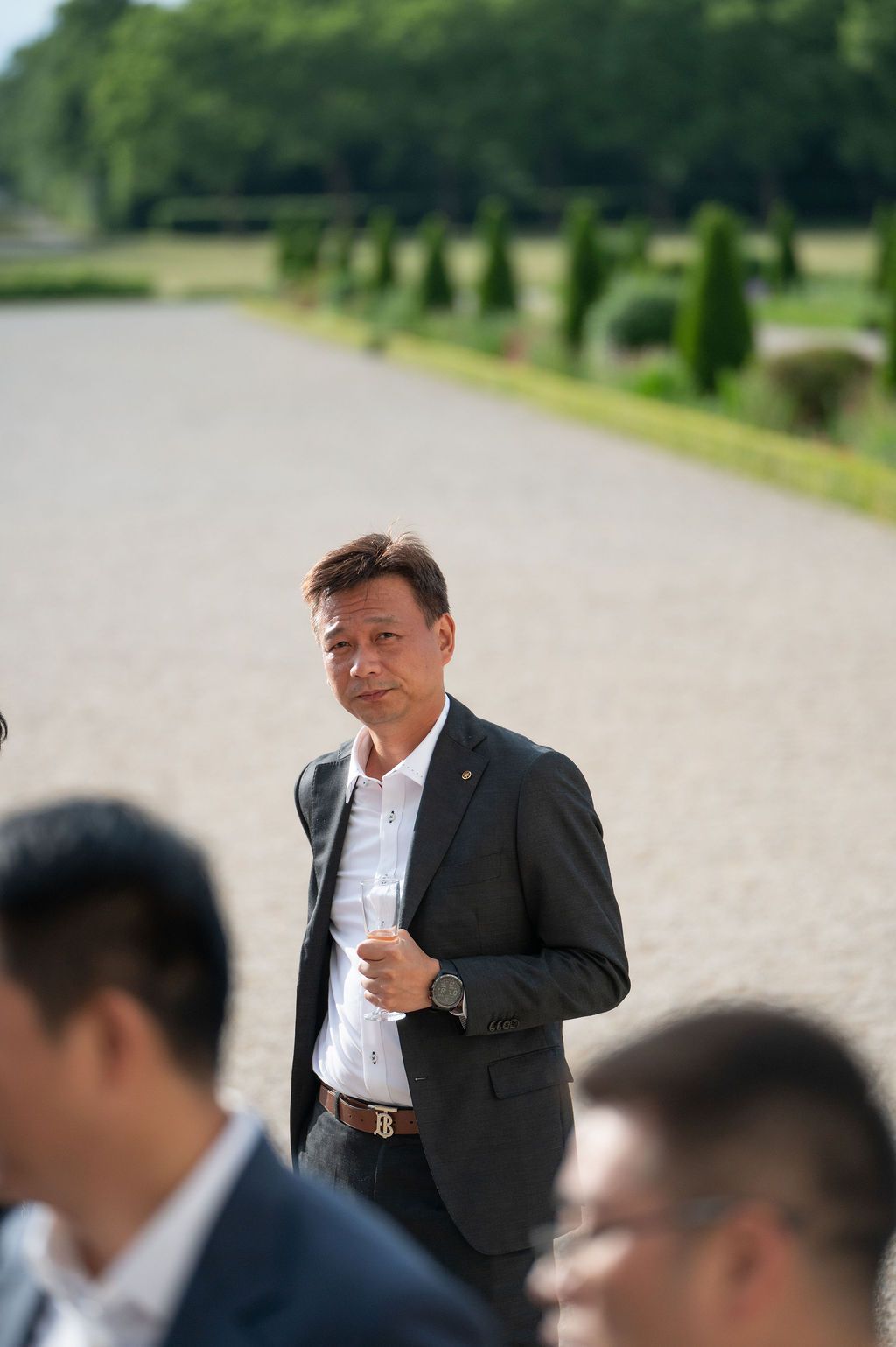 Plan serré sur un cadre souriant tenant son verre, allée gravillonnée du domaine.