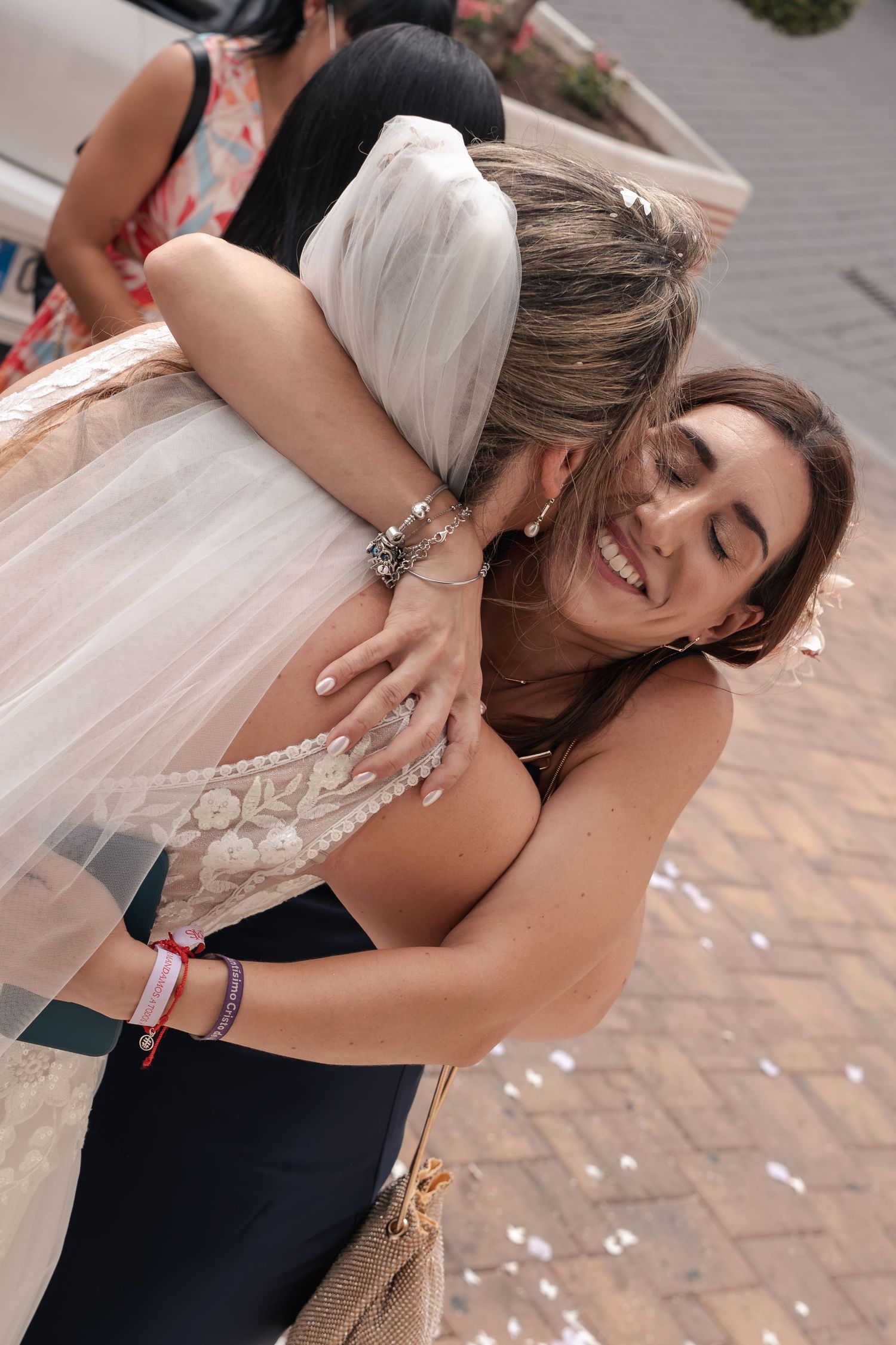 A smiling woman in a navy dress hugs a bride in a lace wedding gown and veil on a brick path scattered with confetti.