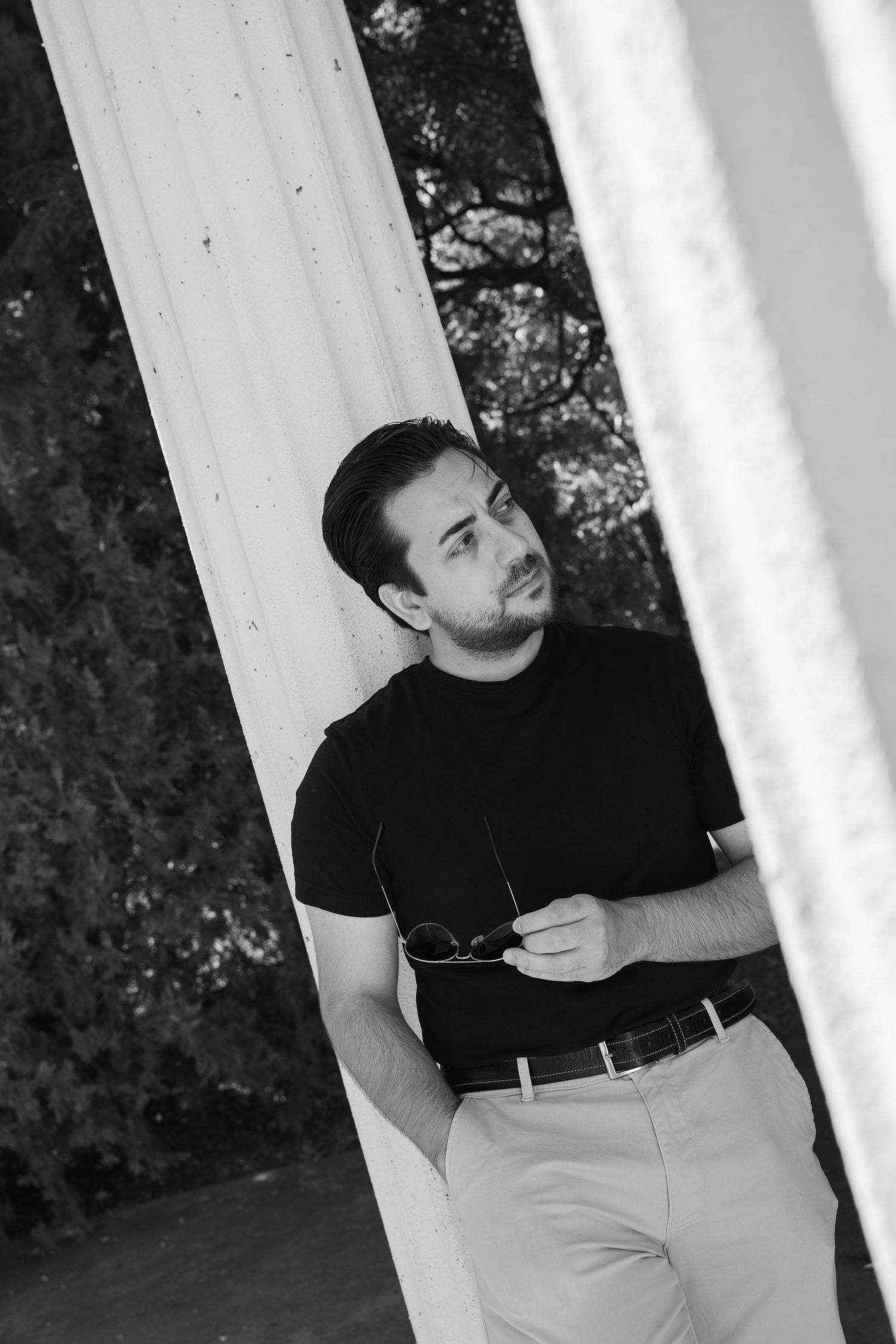 Black and white portrait of a bearded man in a dark shirt leaning against a white column, holding sunglasses and gazing away.