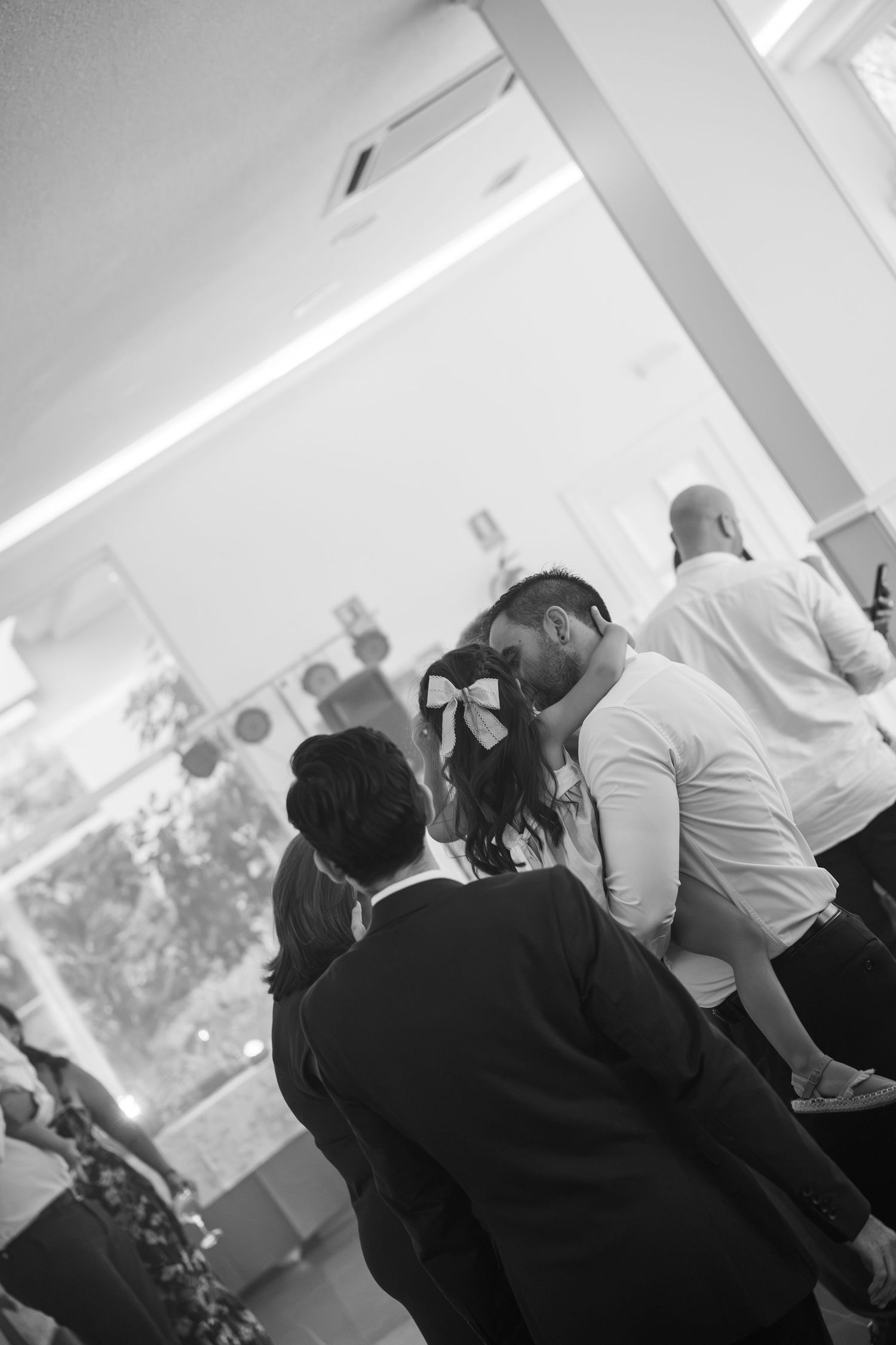 Black and white photo of a man in a white shirt kissing a woman with a bow in her hair at a lively indoor wedding celebration.
