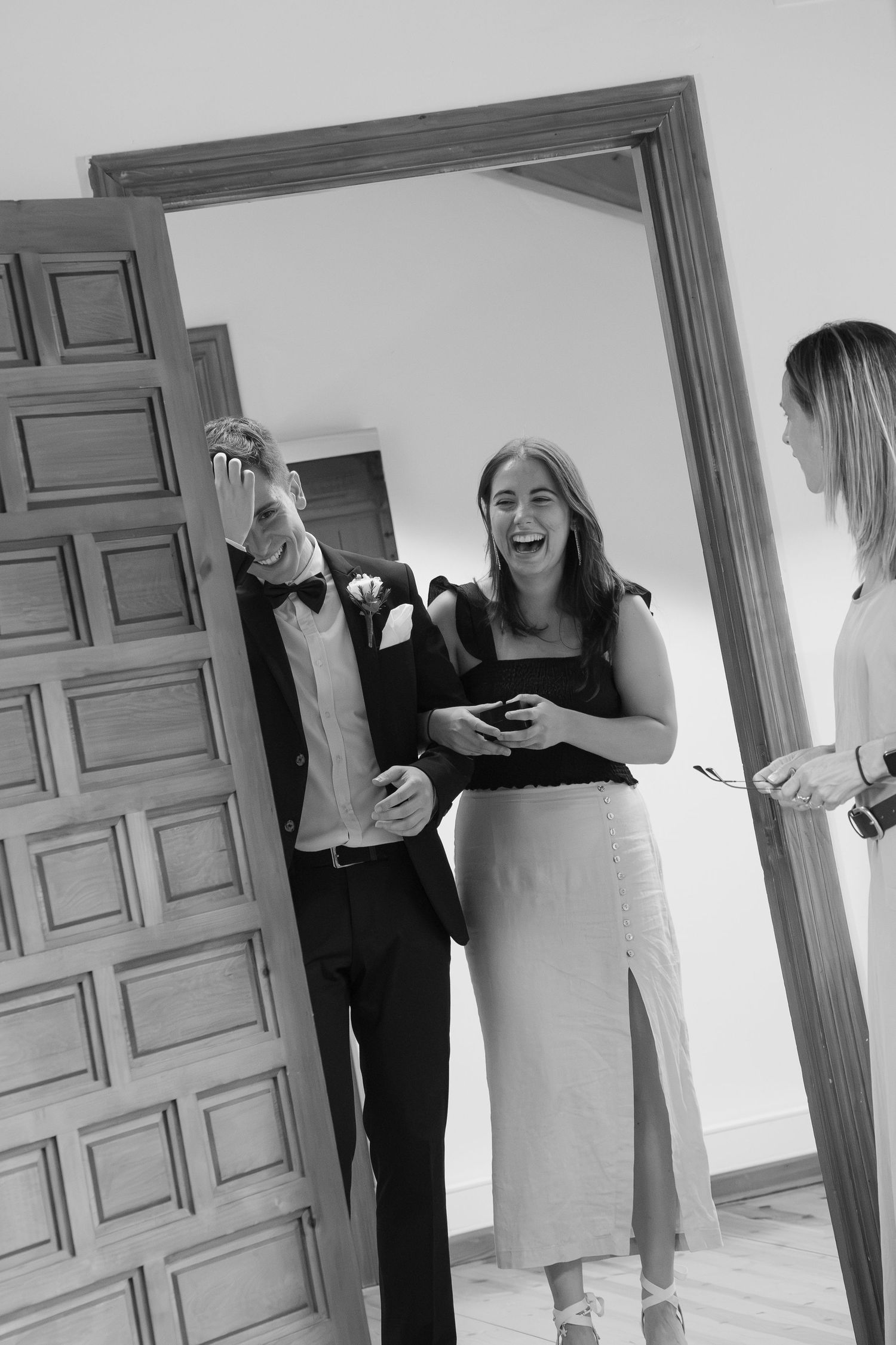 Black and white photo of a laughing woman in a black top and skirt beside a man in a suit and bow tie shielding his face near a wooden door.