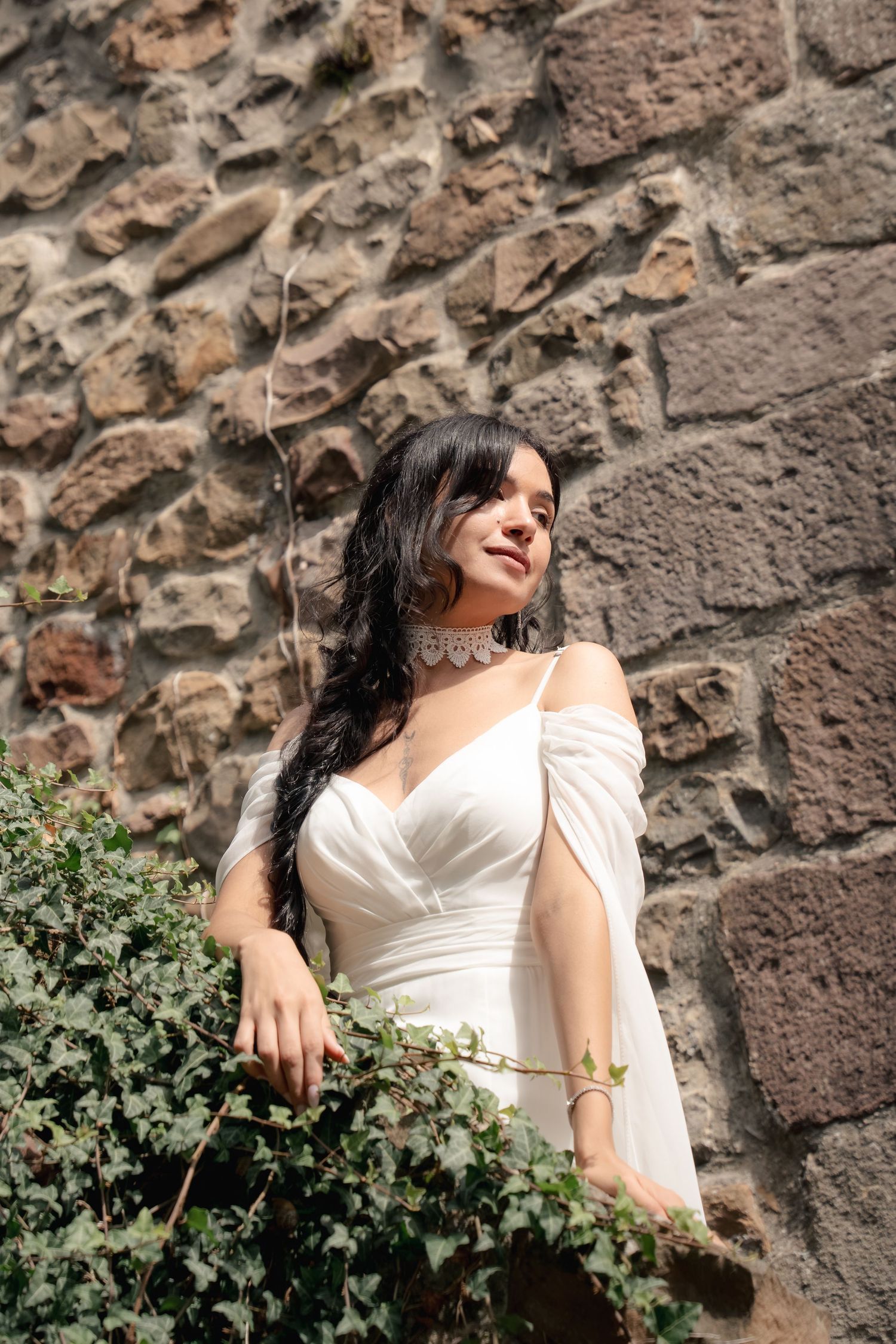 Woman with long dark hair in a white draped wedding dress and lace choker stands beside green ivy against a rustic stone wall.