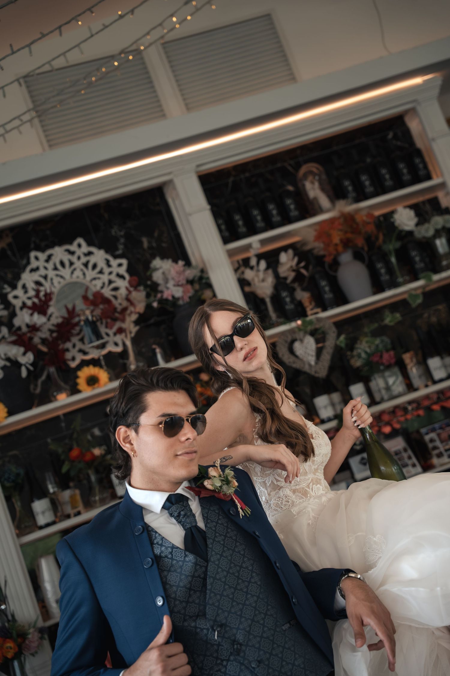 Stylish couple in sunglasses, the groom in a navy suit and the bride in a lace wedding gown, posing in a floral shop with wine bottles.