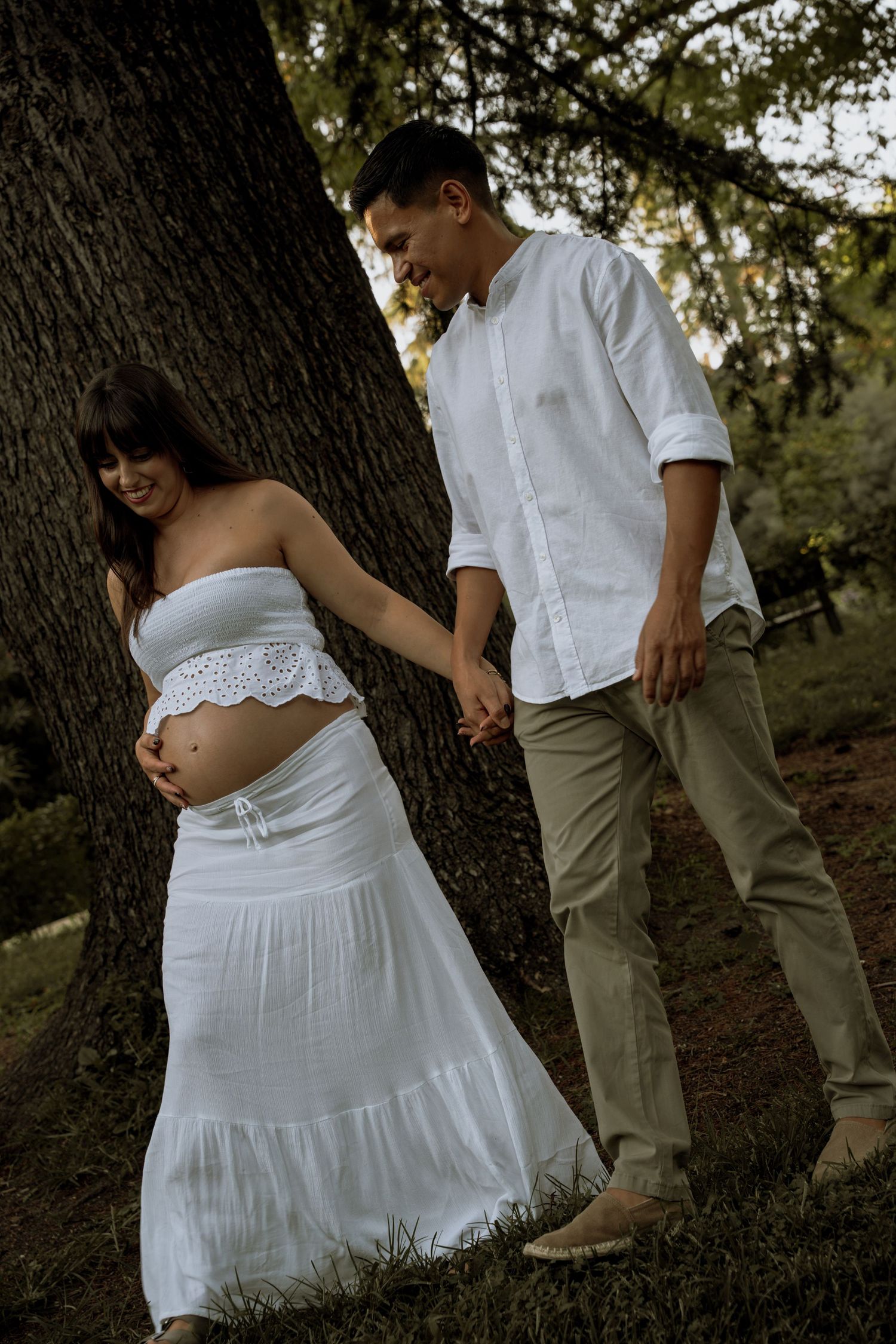 Expecting couple in white outfits holding hands and walking beside a large tree during an outdoor maternity photoshoot.