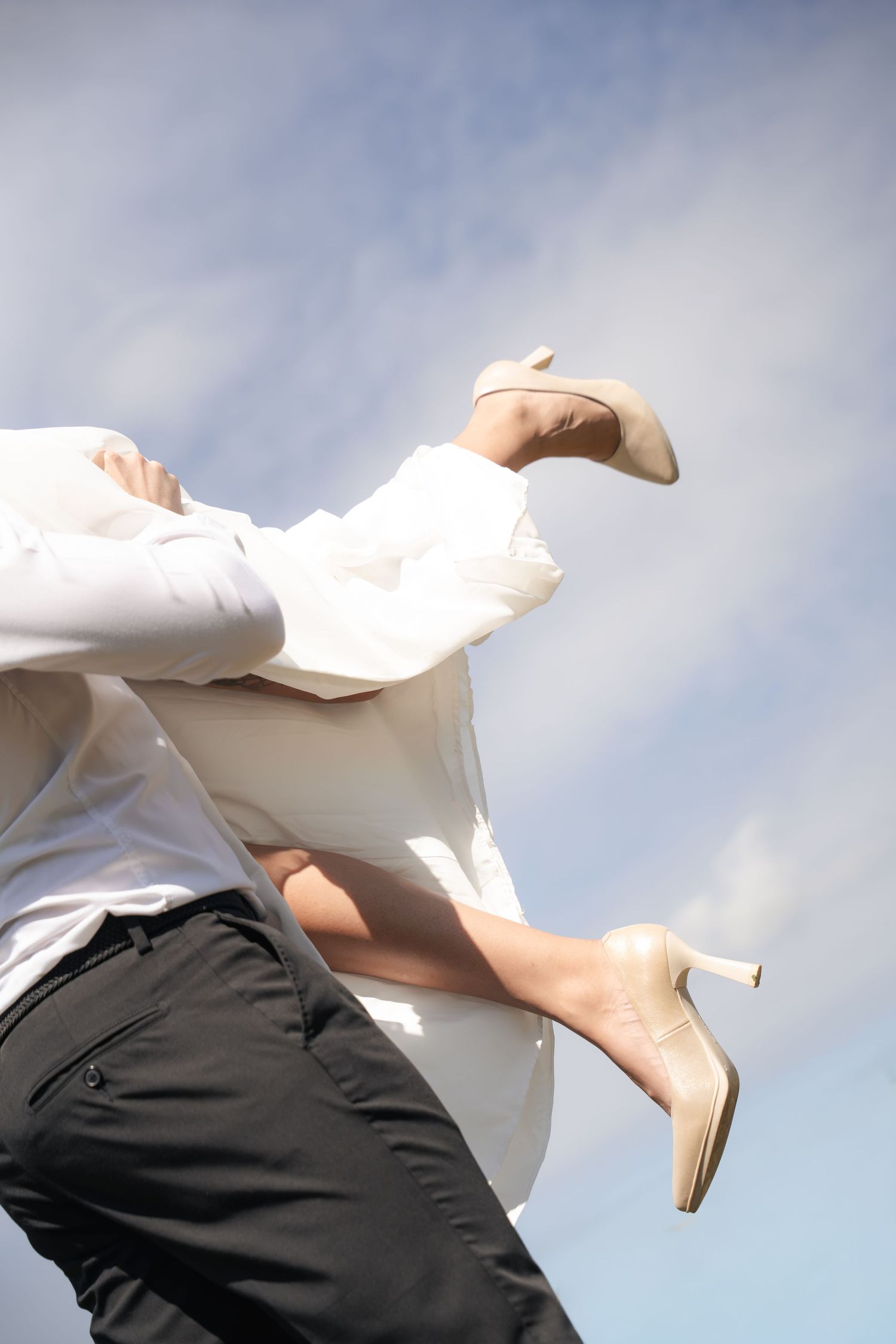 Person in white shirt lifting a woman in beige high heels mid-air against a bright blue sky with soft clouds.