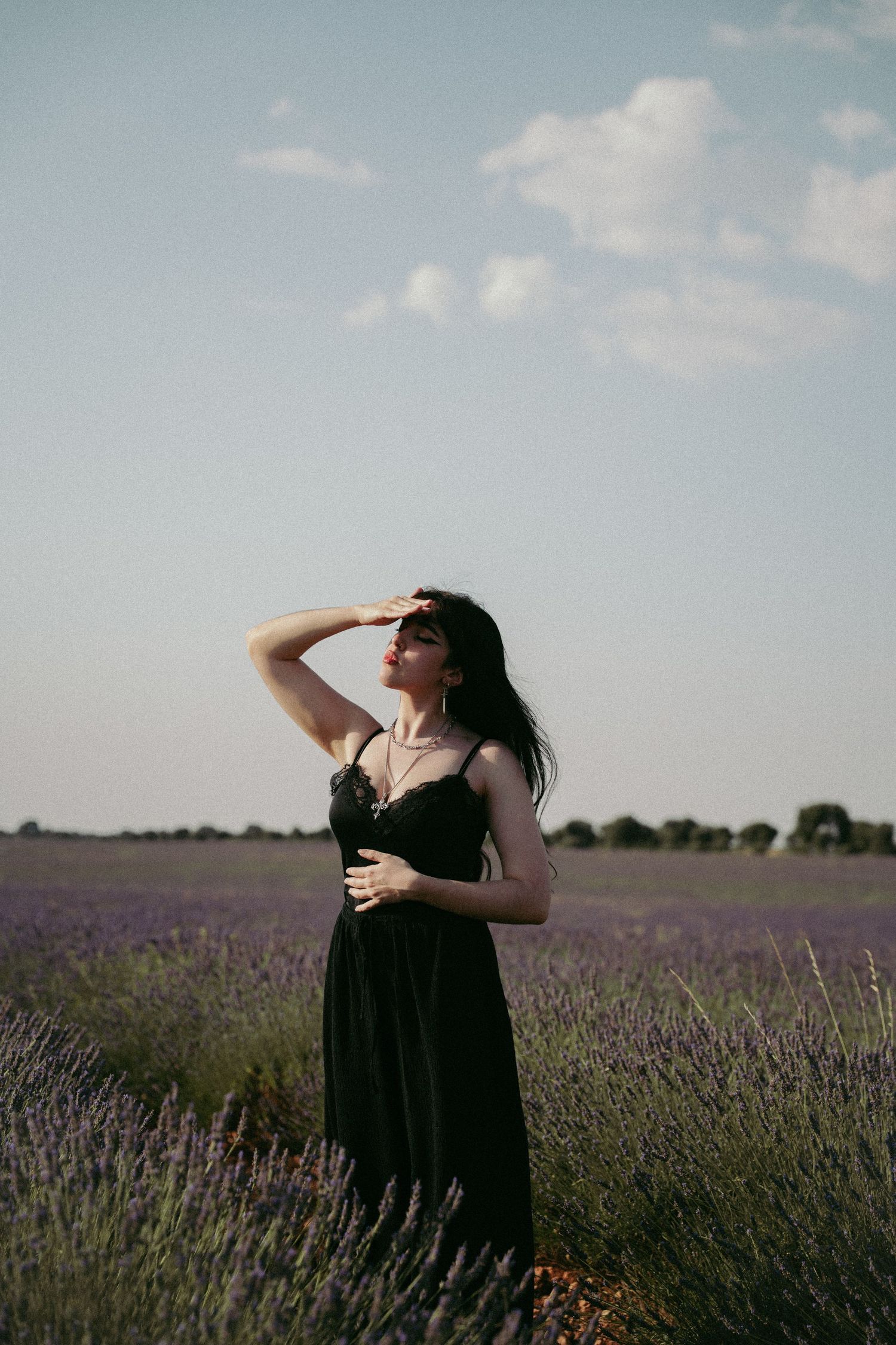 Woman in a black dress shields her eyes from the sun while standing in a blooming lavender field under a soft blue sky.