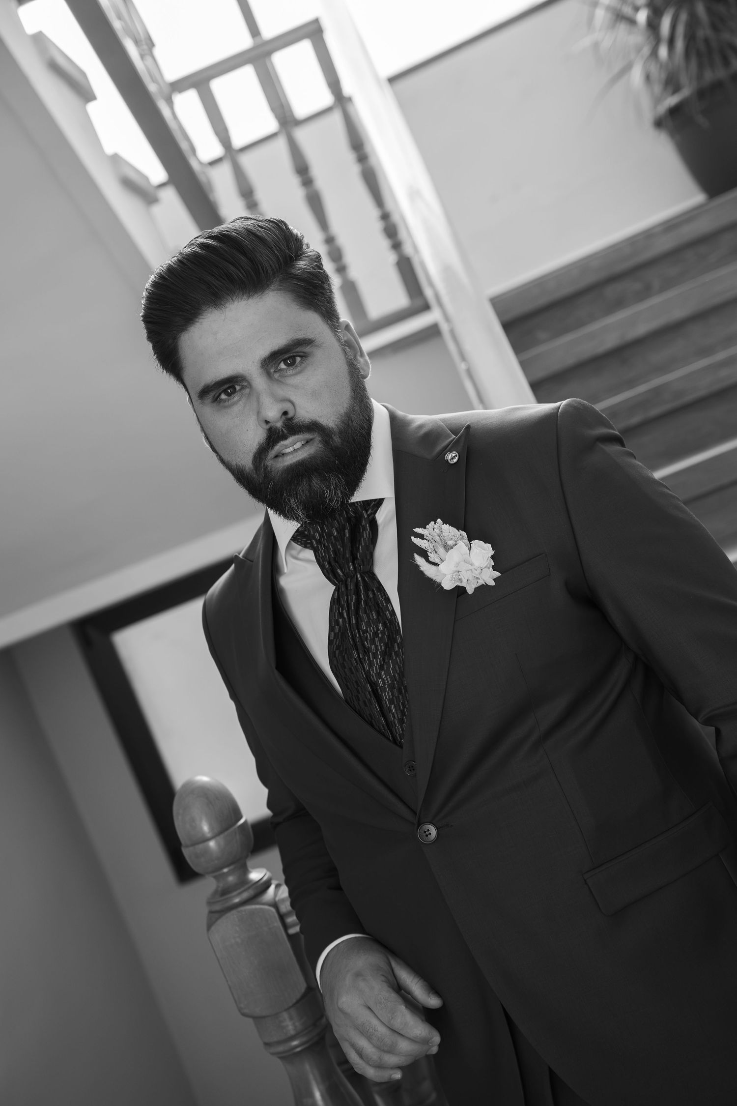 Black and white portrait of a bearded groom in a dark three-piece suit with patterned tie and floral boutonniere on a staircase.