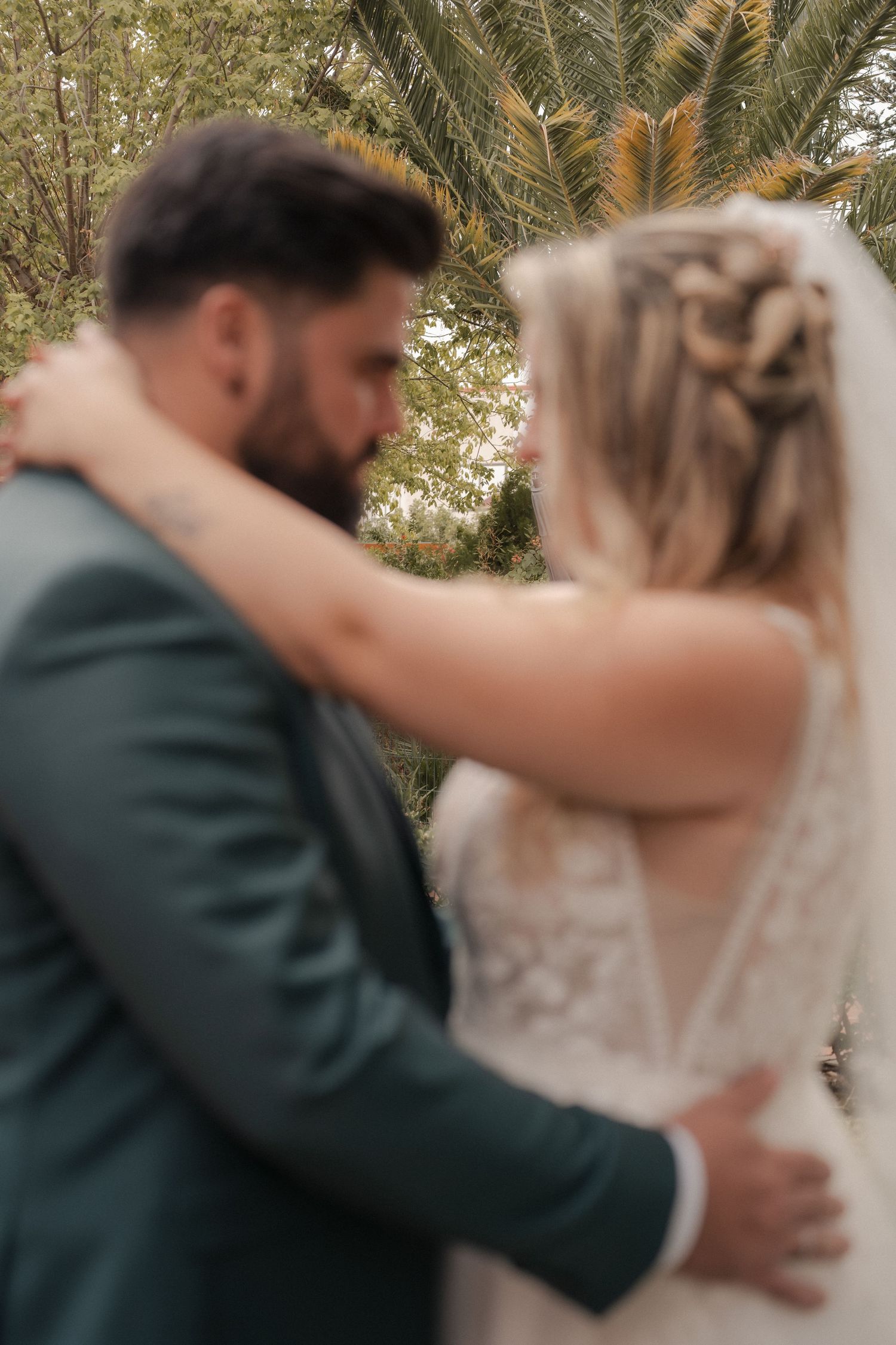 Bride in a lace wedding gown and groom in a green suit embrace in a softly blurred outdoor garden with palm trees.