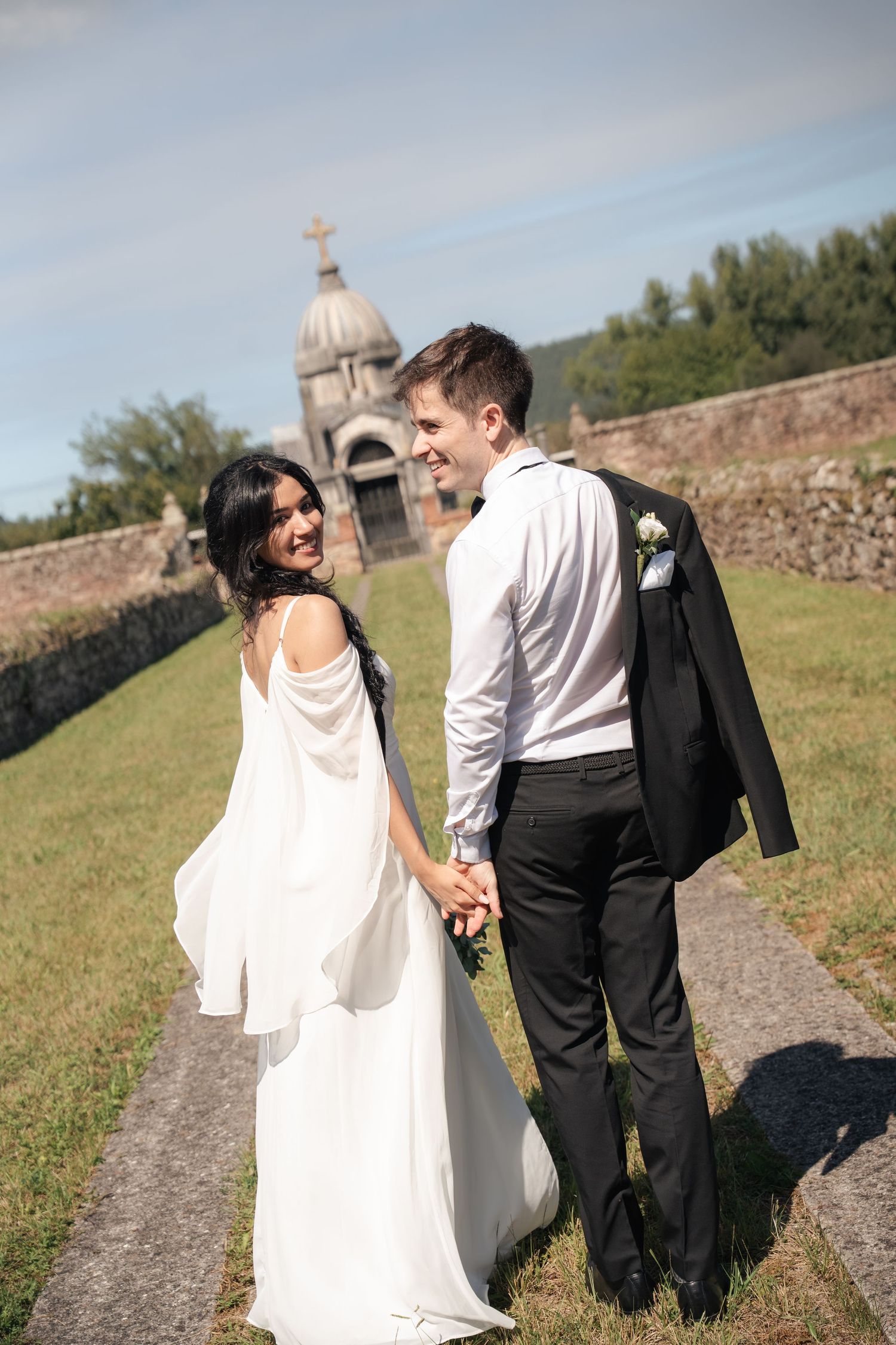 Smiling newlywed couple holding hands walking outdoors near a historic stone chapel on their wedding day.