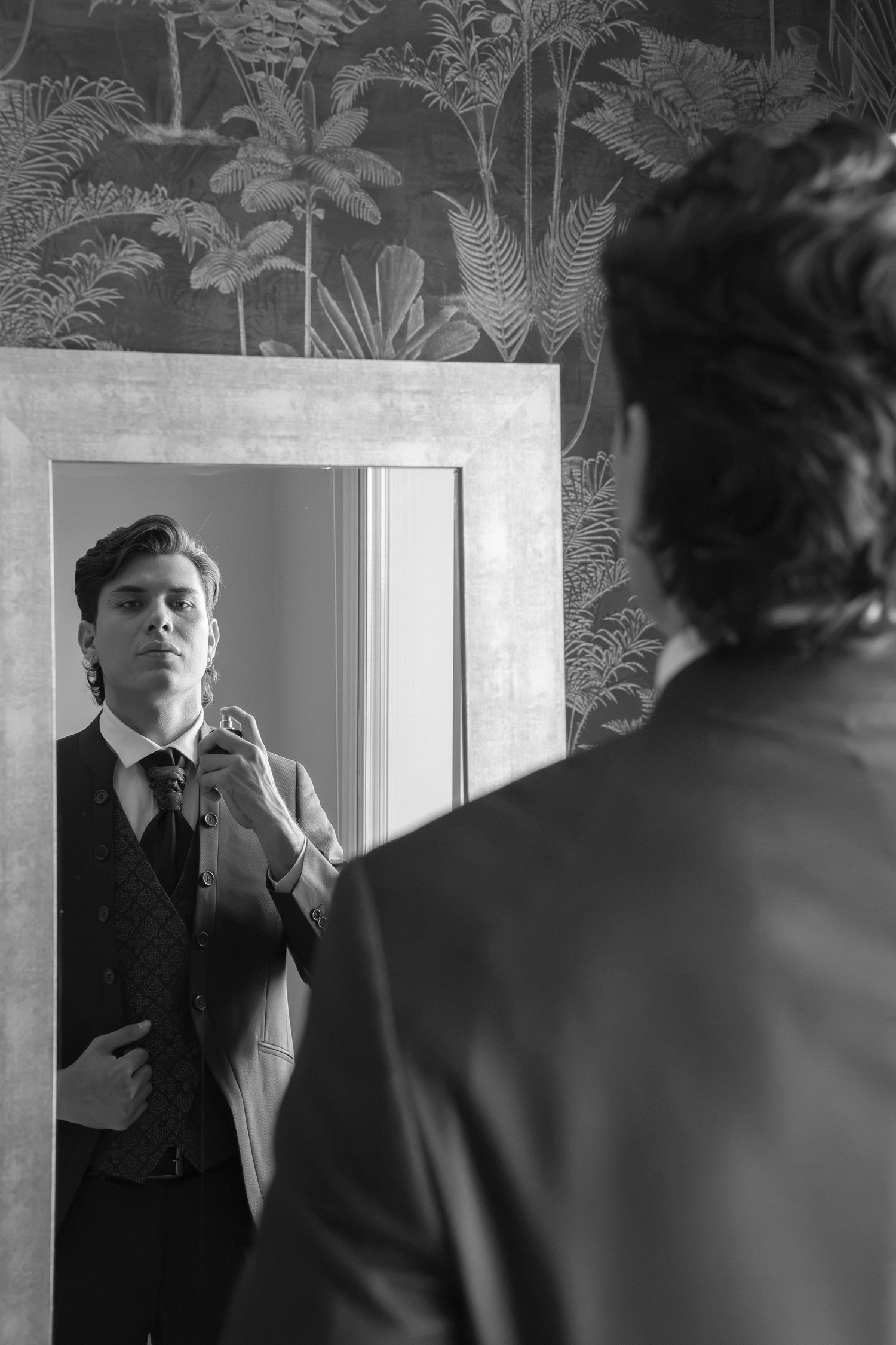 Groom adjusting his tie while looking in an ornate mirror in black and white wedding preparation photo.