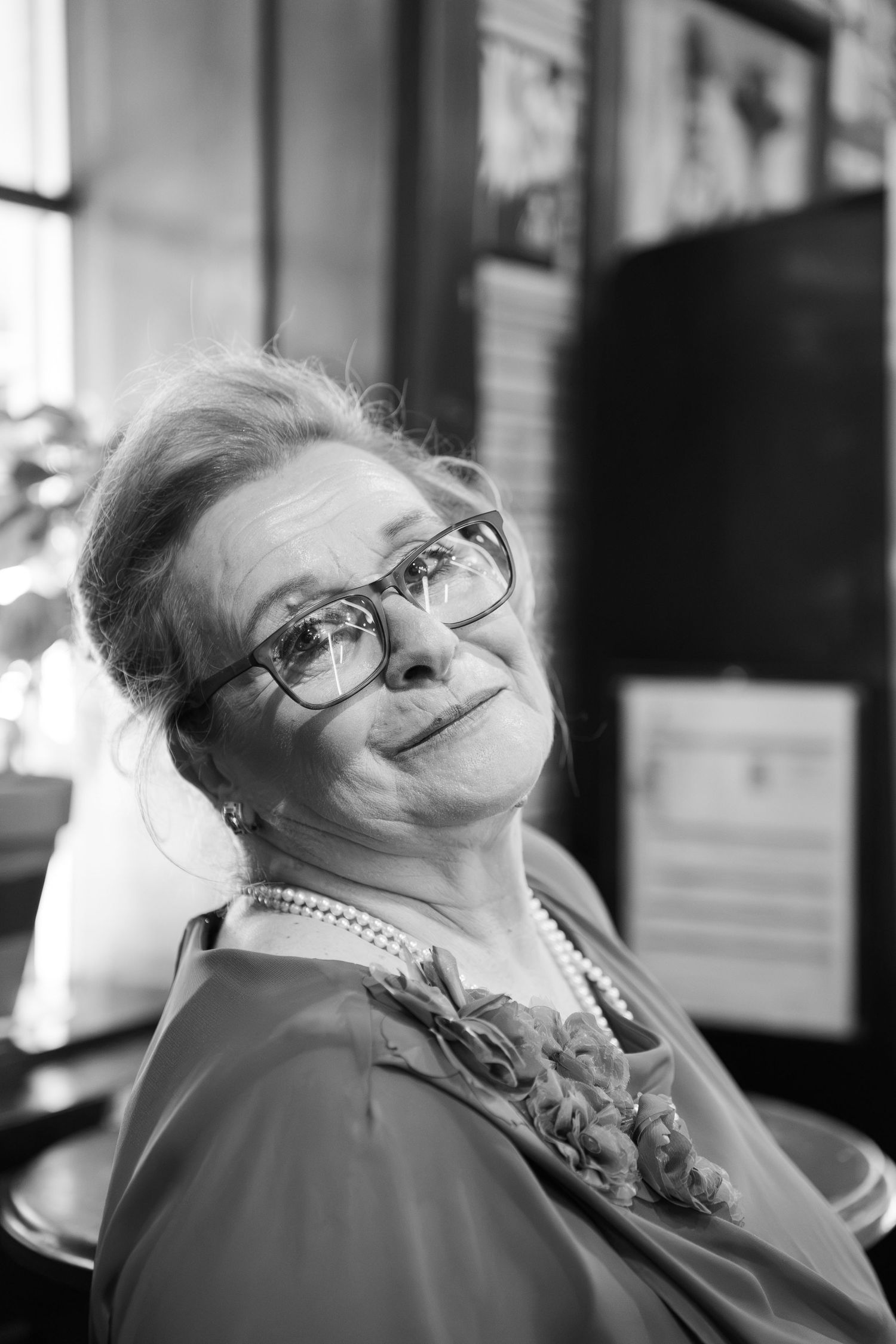 Black and white portrait of a smiling older woman with glasses, pearl necklace and a floral corsage, tilting her head in a cozy room.