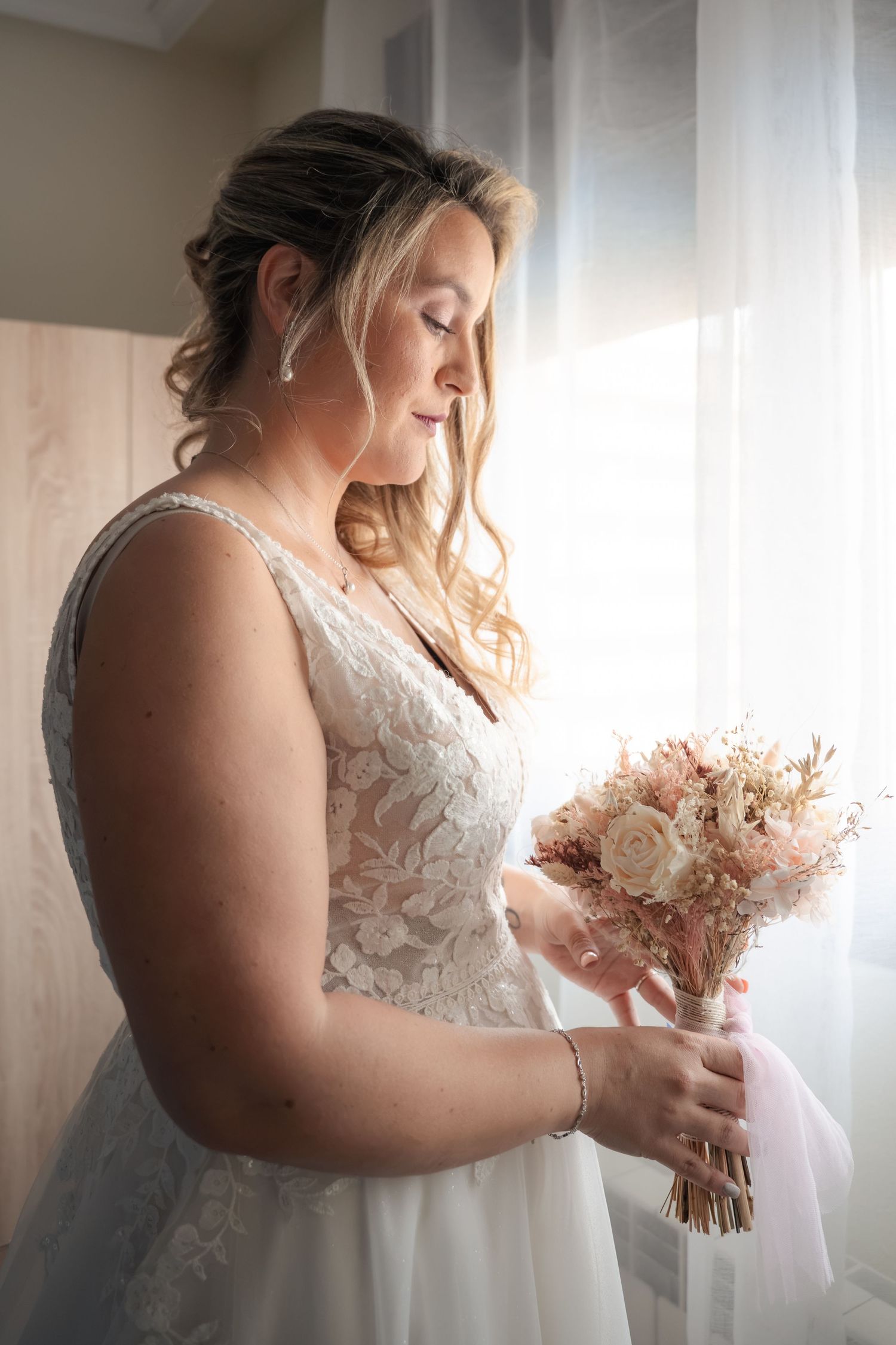 Bride in a lace wedding gown holds a dried-flower bouquet with pink ribbon while gazing down beside a softly lit window.