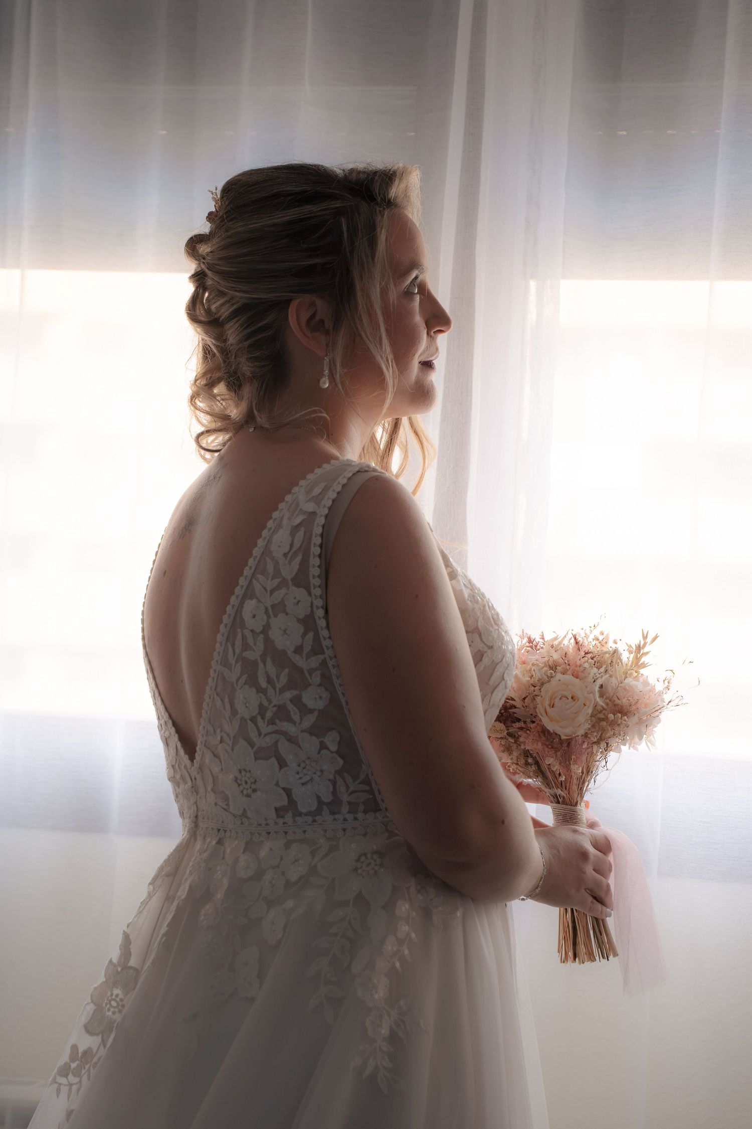 Bride in a lace wedding gown with an open back and an updo holds a pink and peach dried flower bouquet while gazing out a sunlit window.